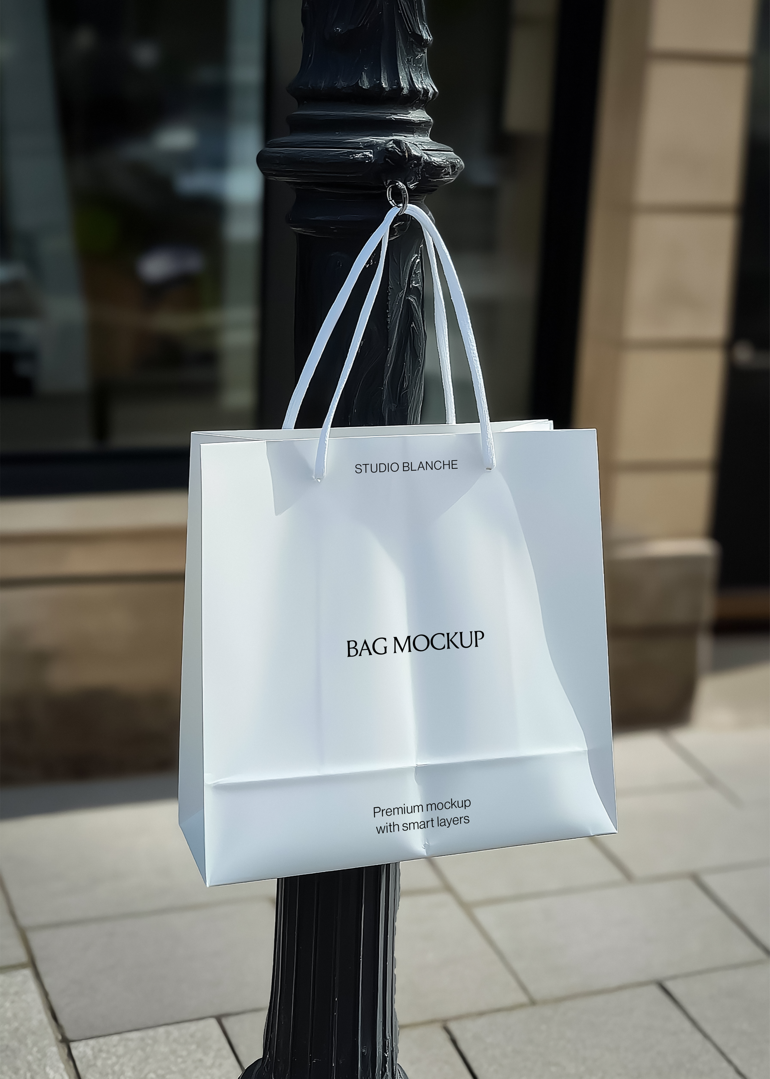 White paper shopping bag mockup with white handles hanging from a black street post, shopfront glass behind in sunlight