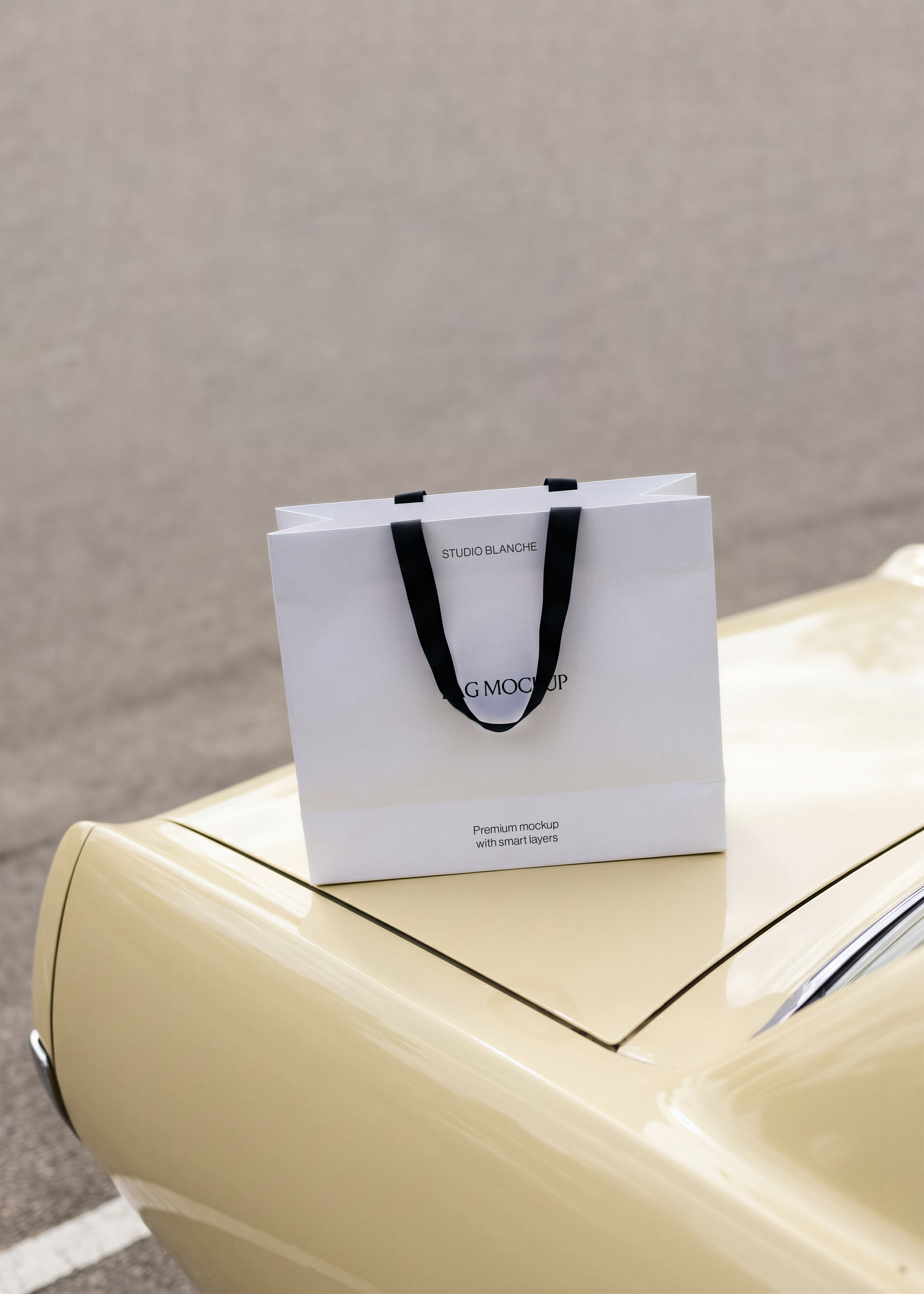 Studio Blanche shopping bag mockup with black handles on a glossy cream vintage car bonnet, tarmac road behind