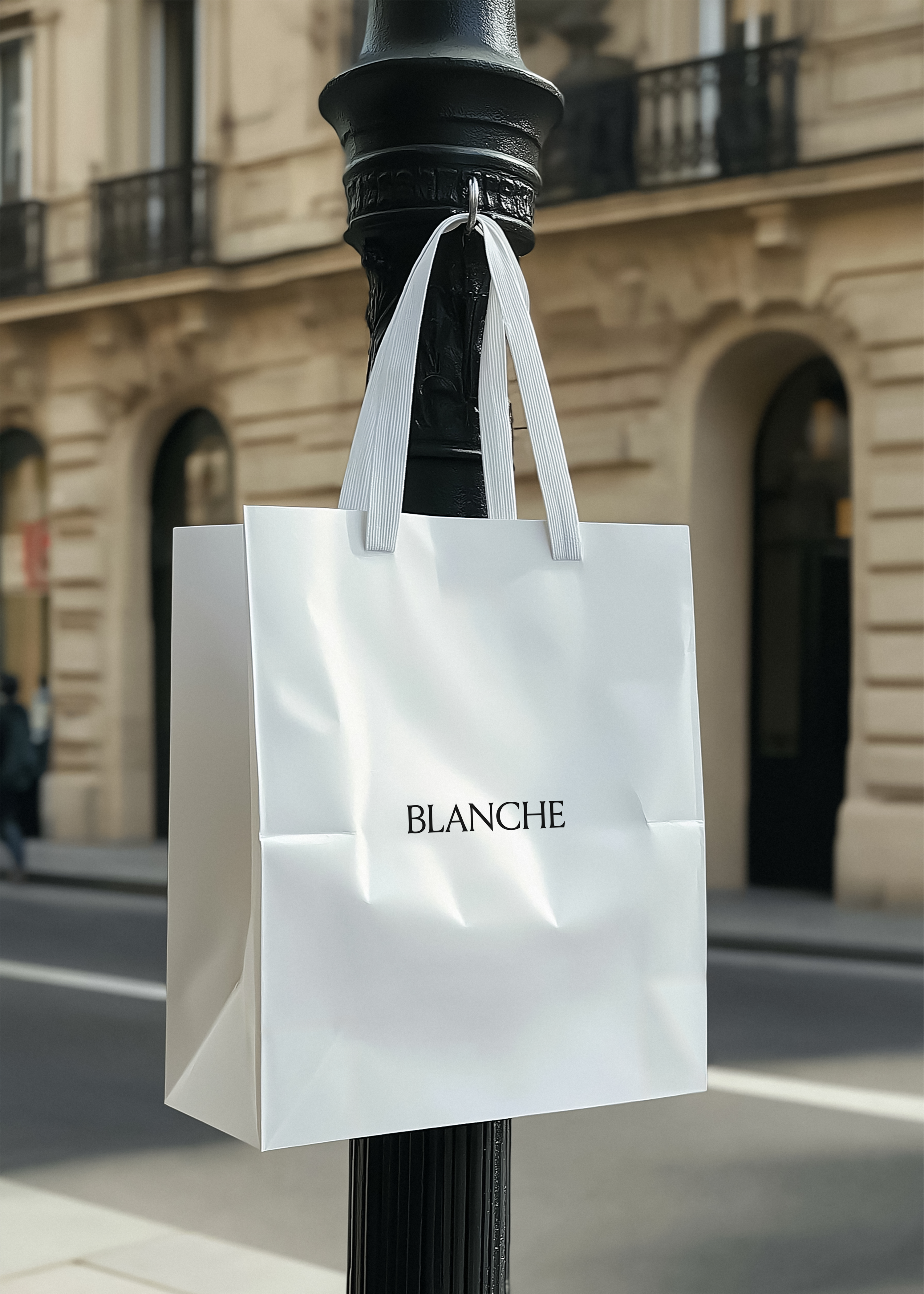 White paper shopping bag with Blanche logo hanging from a black lamp post before a Parisian Haussmann facade