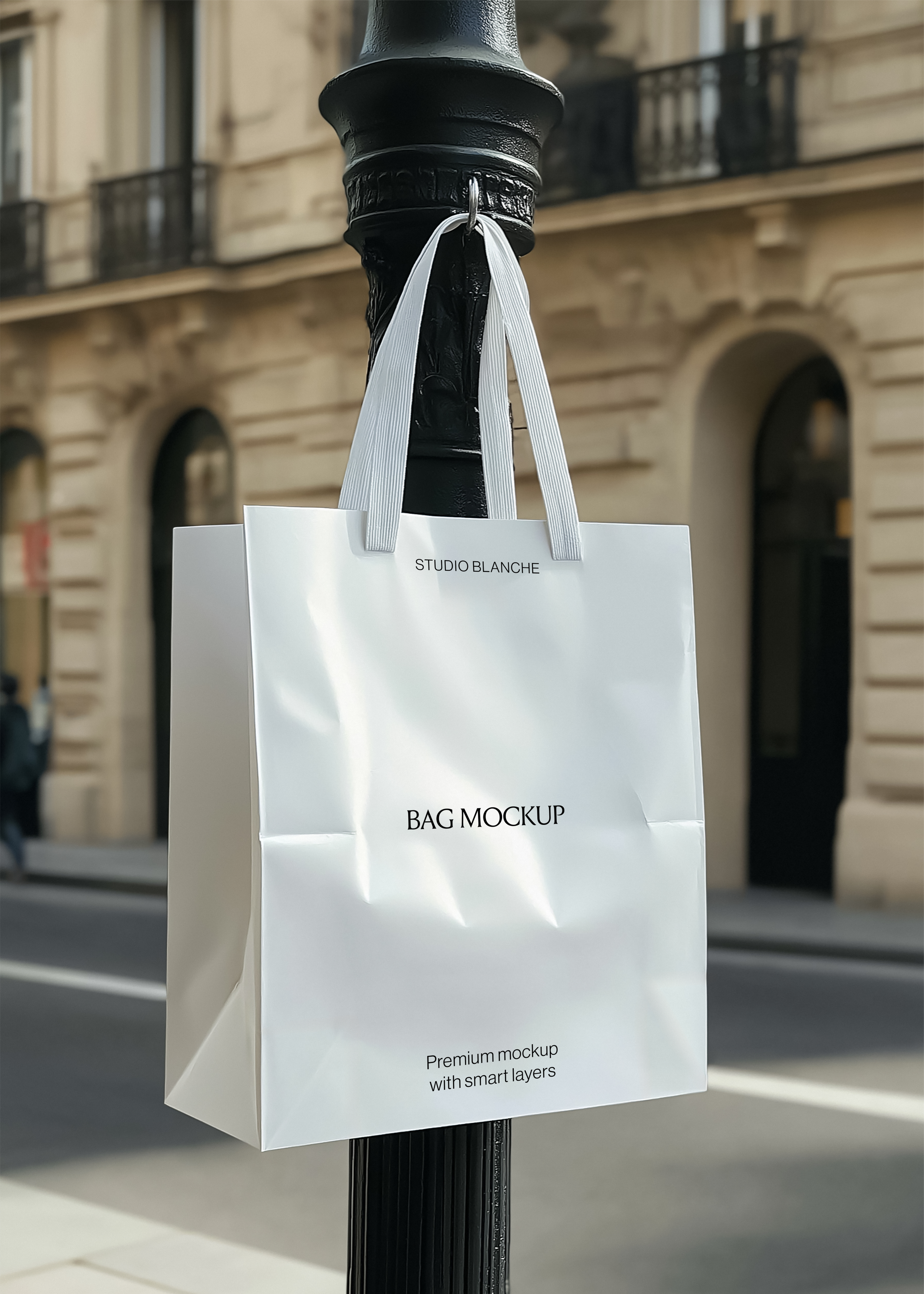 White paper shopping bag mockup with white handles on a black lamp post, blurred Haussmann building and street behind