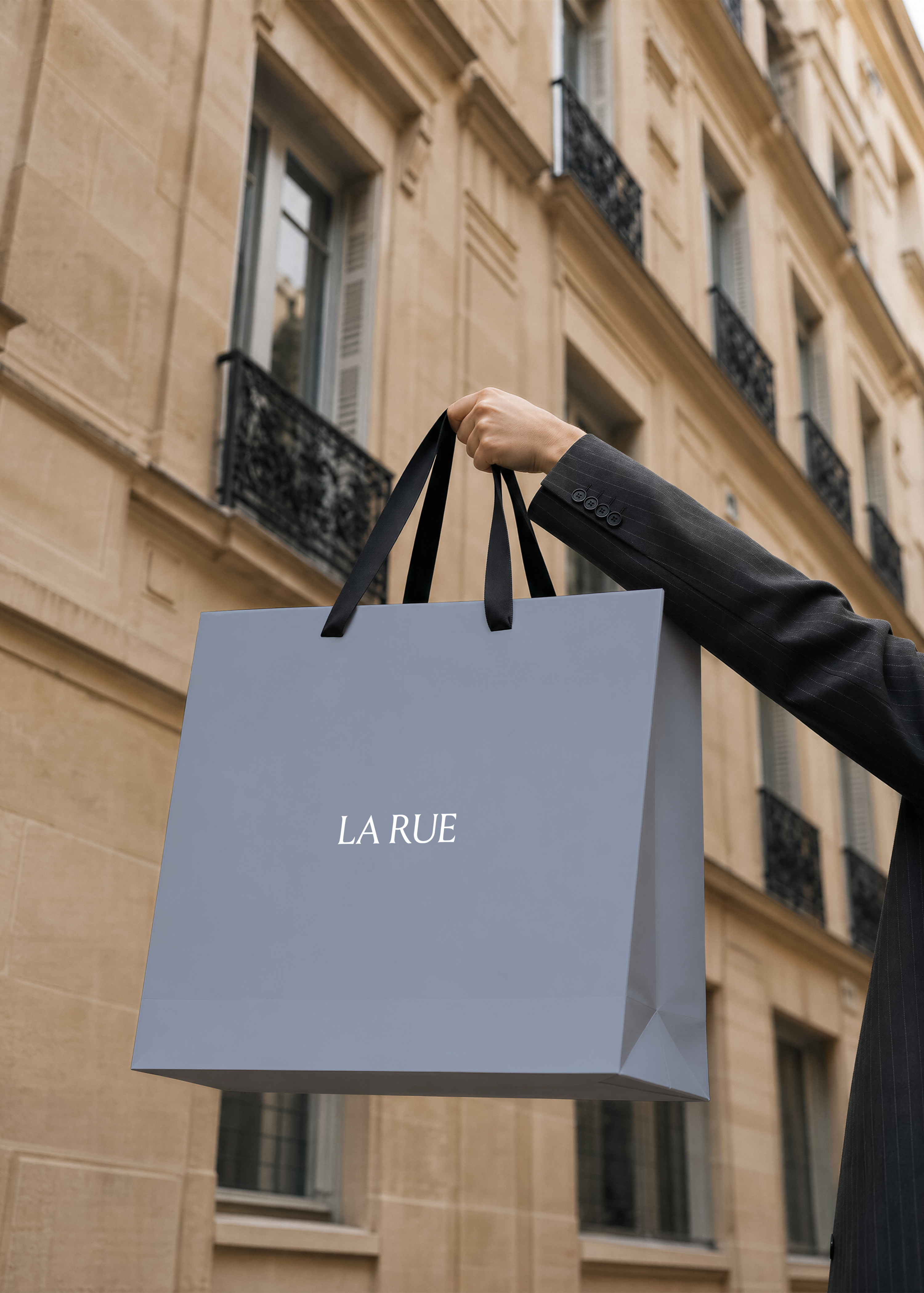 Hand in a dark pinstripe suit holding a pale grey LA RUE shopping bag with black handles against a Parisian stone facade