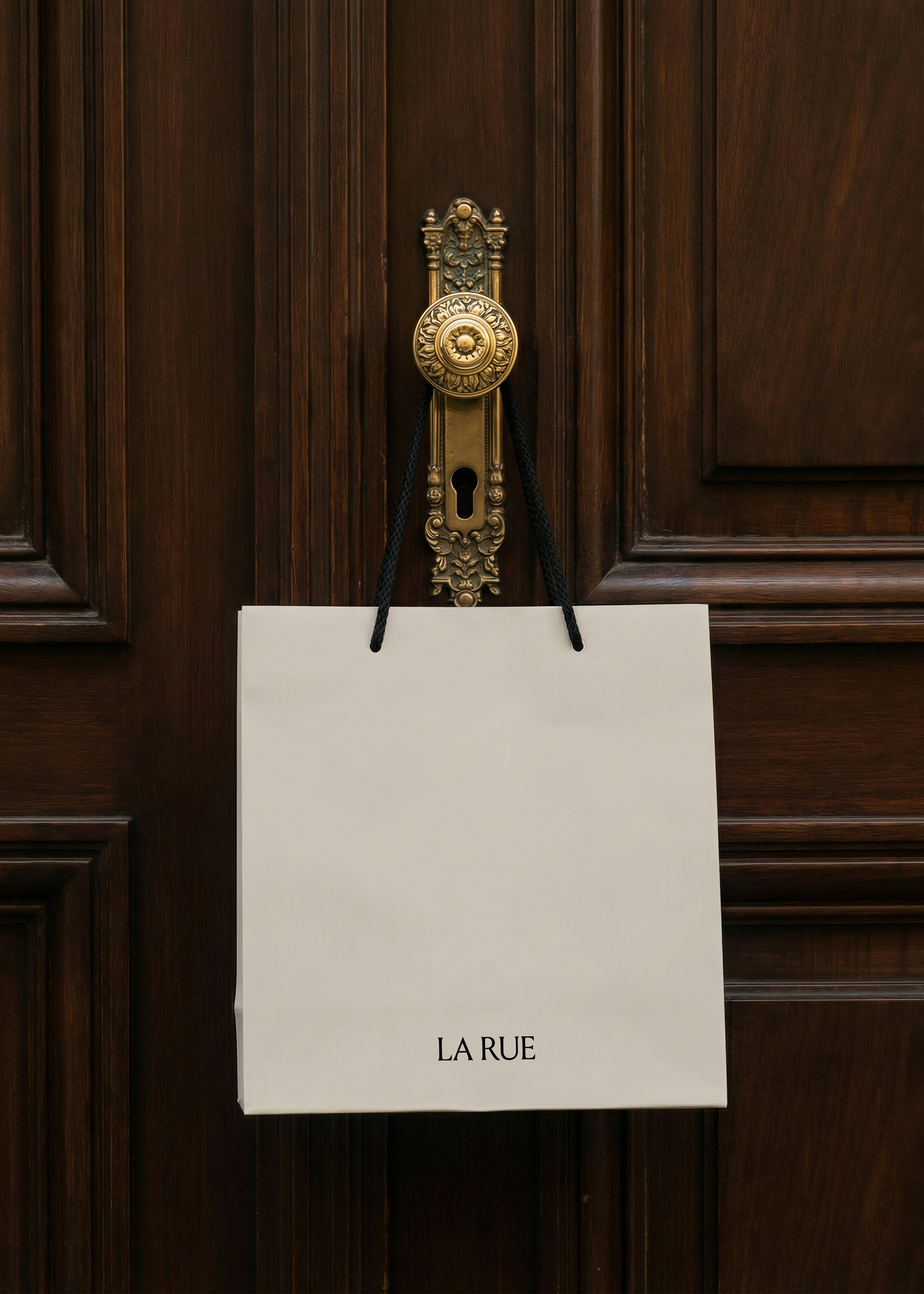 Cream shopping bag mockup with black rope handles hanging from an ornate brass door knob on dark panelled wood door