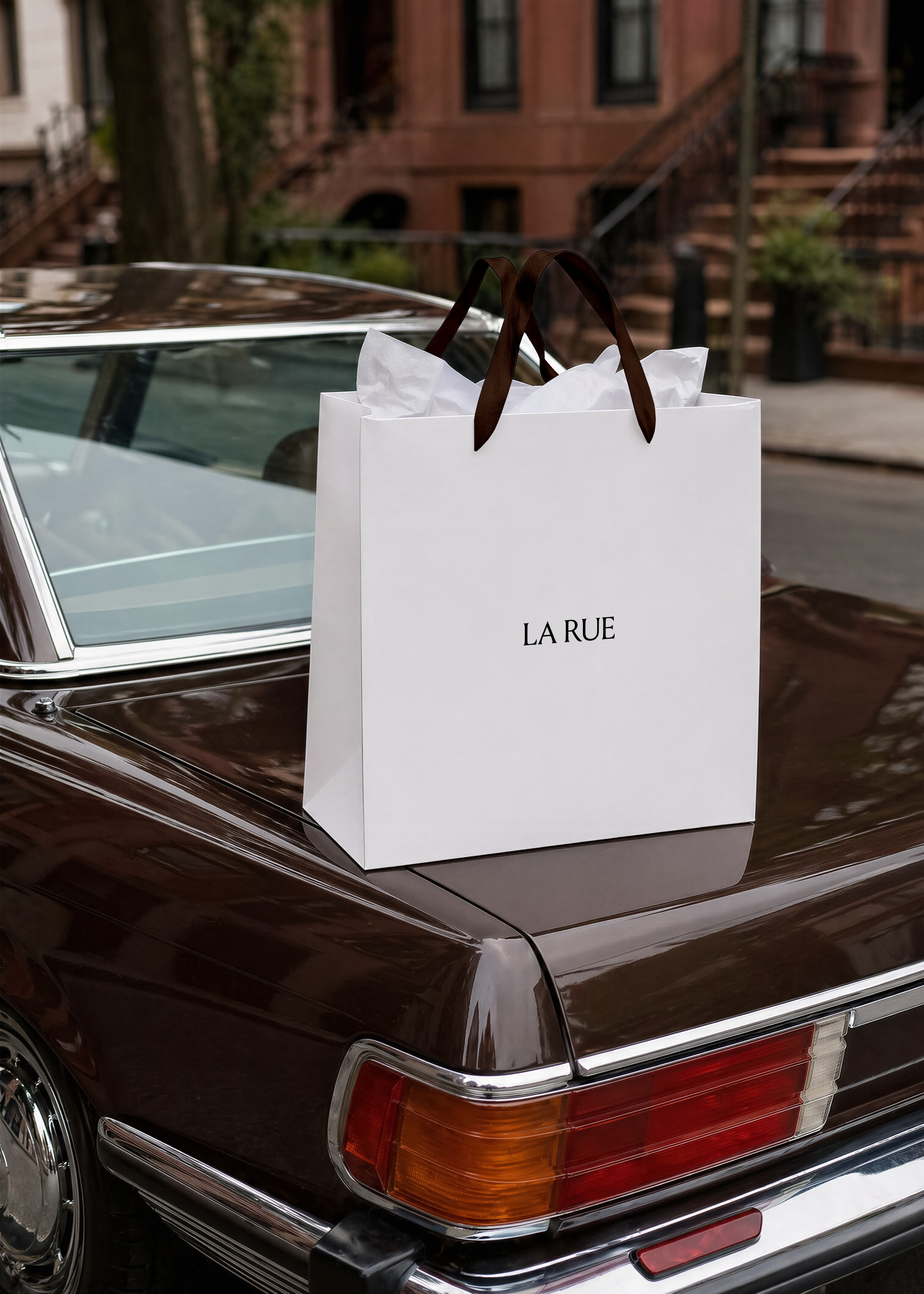 White shopping bag mockup with brown ribbon handles and tissue paper on burgundy vintage car boot, brownstone street
