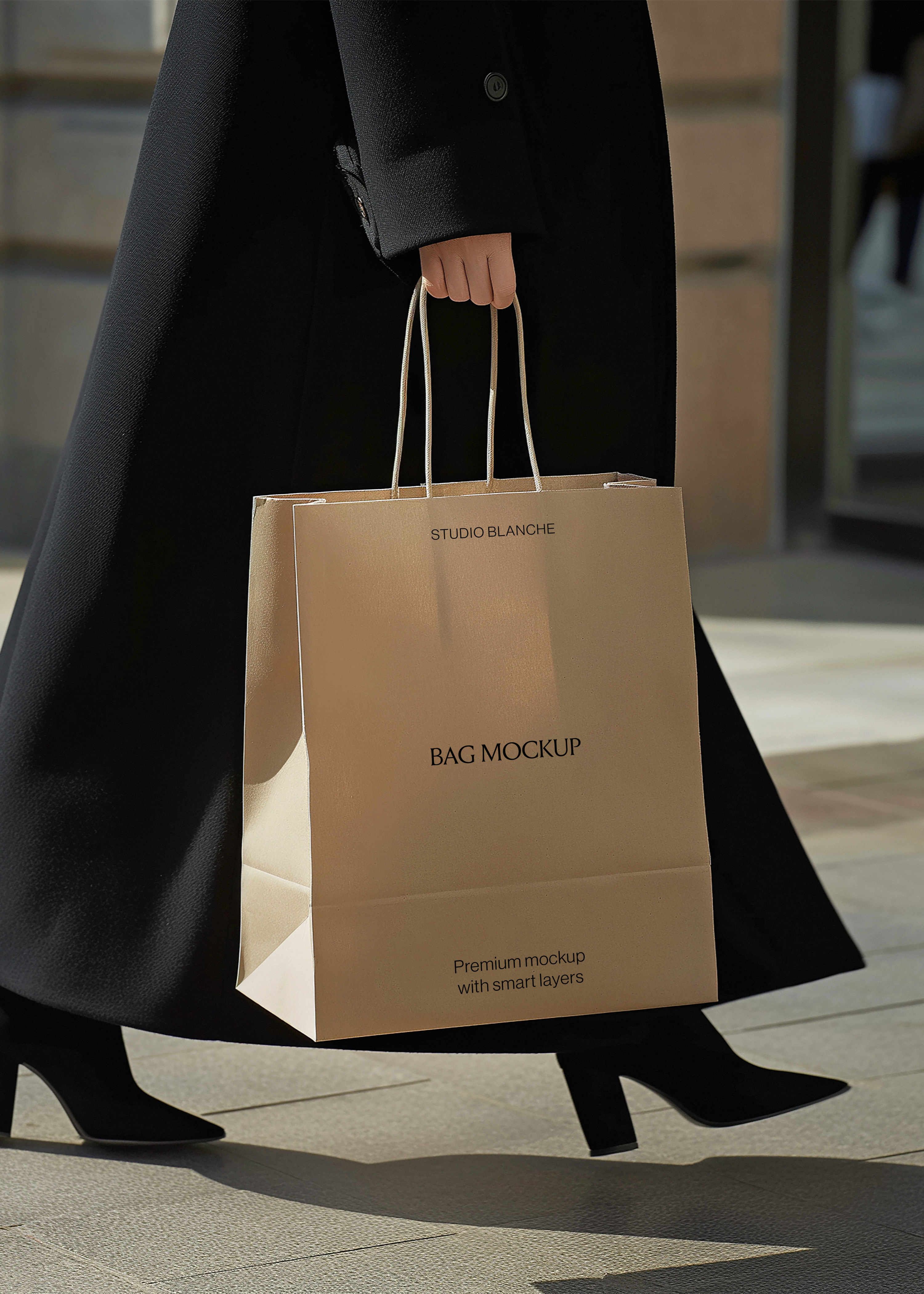Tan paper shopping bag mockup with twisted handles held in one hand beside a long black coat, sunlit pavement