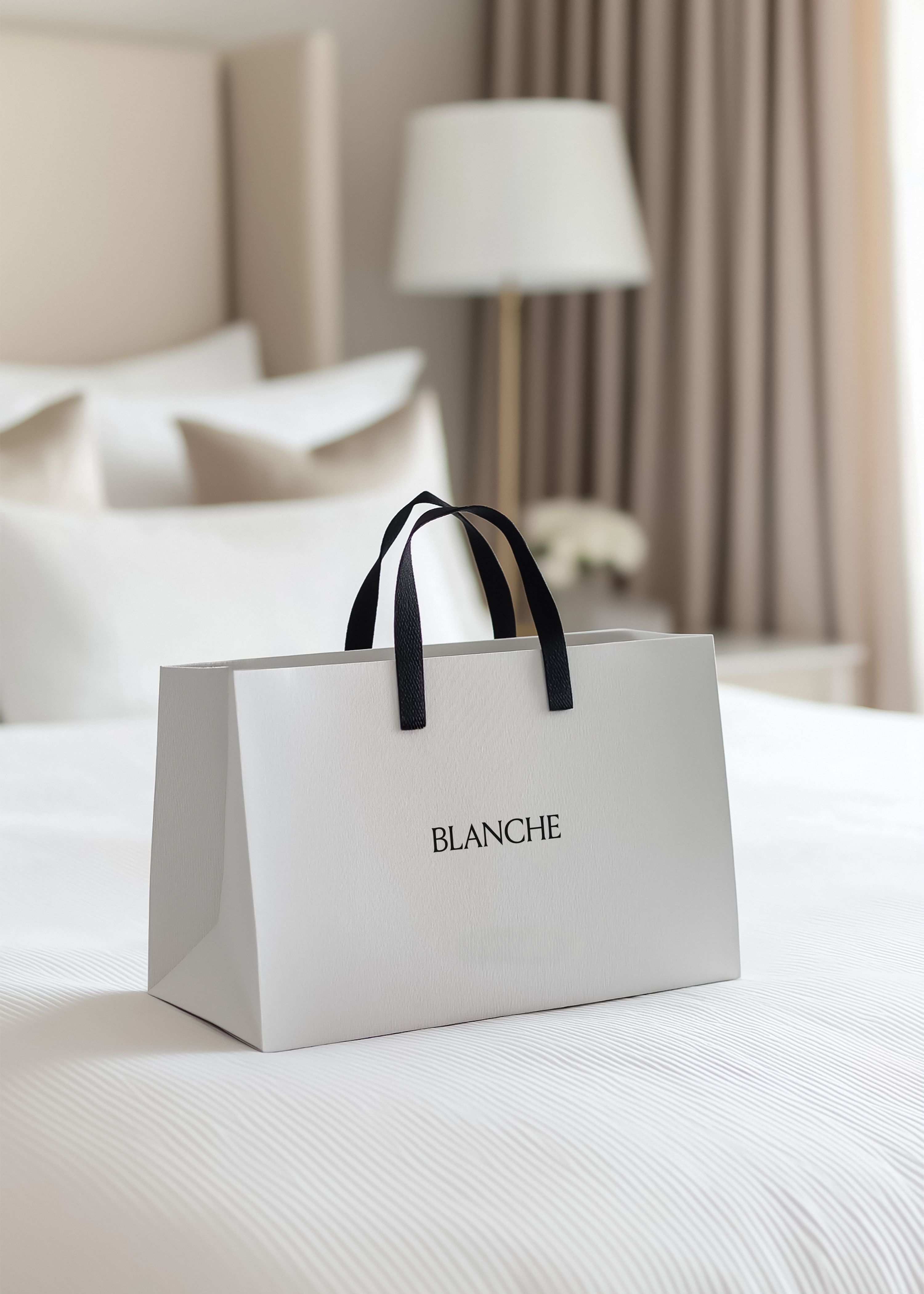 White paper shopping bag with black handles and Blanche logo on a made white bed in a warm hotel suite