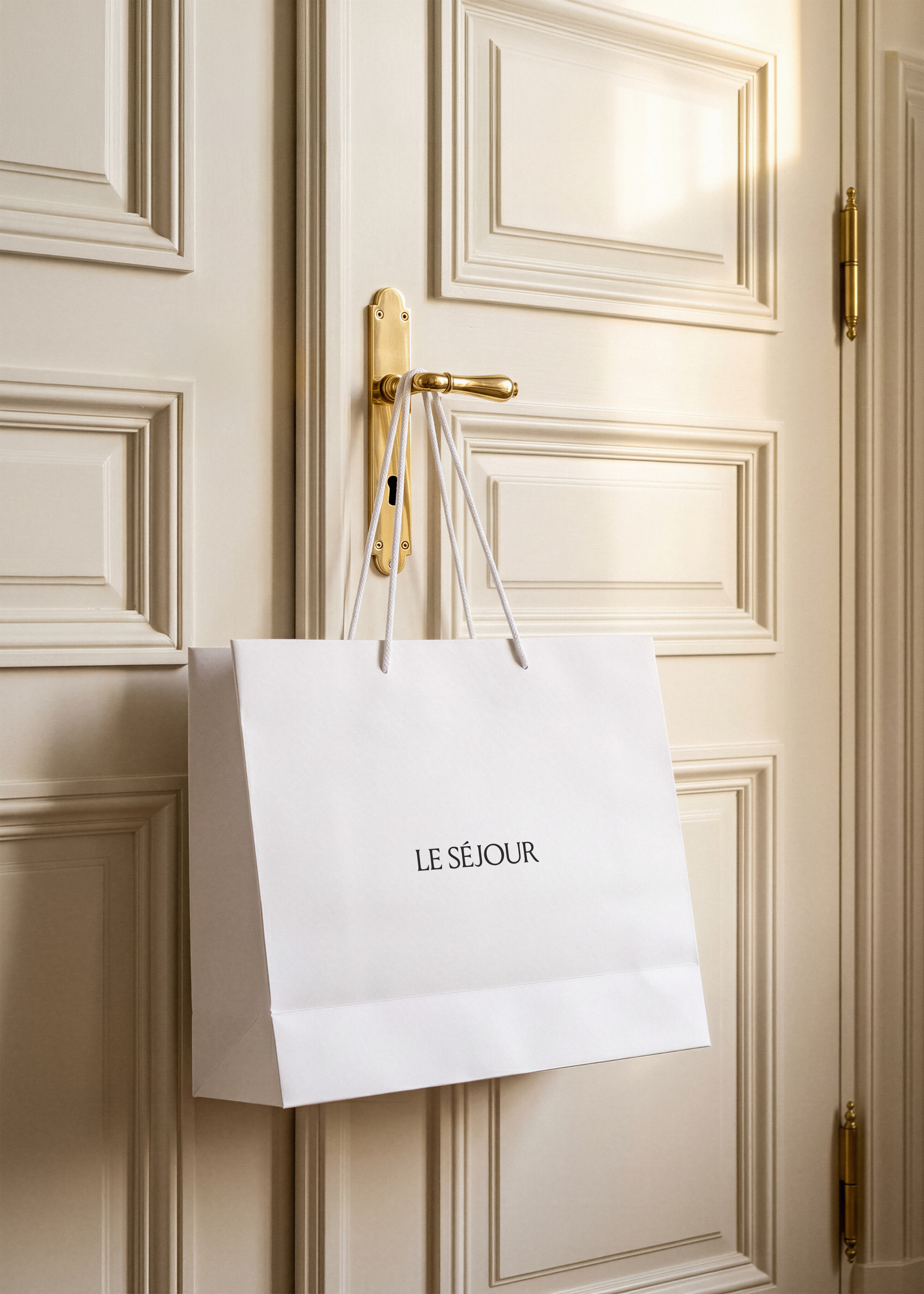 White paper shopping bag mockup hanging from a brass door handle on a cream panelled Parisian door in warm sunlight