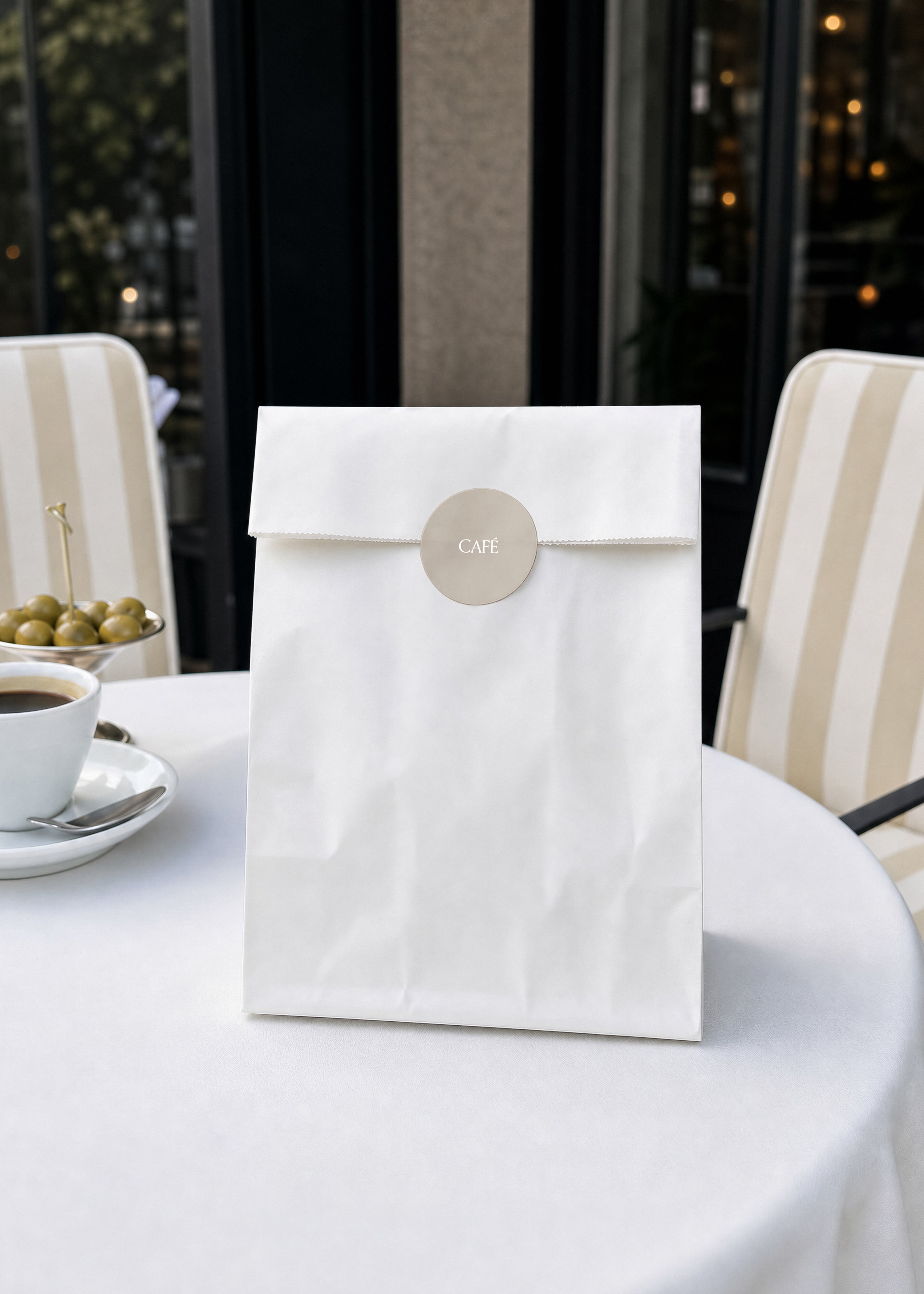 Paper bag mockup with round beige CAFÉ sticker on white tablecloth beside coffee cup and green olives, café terrace