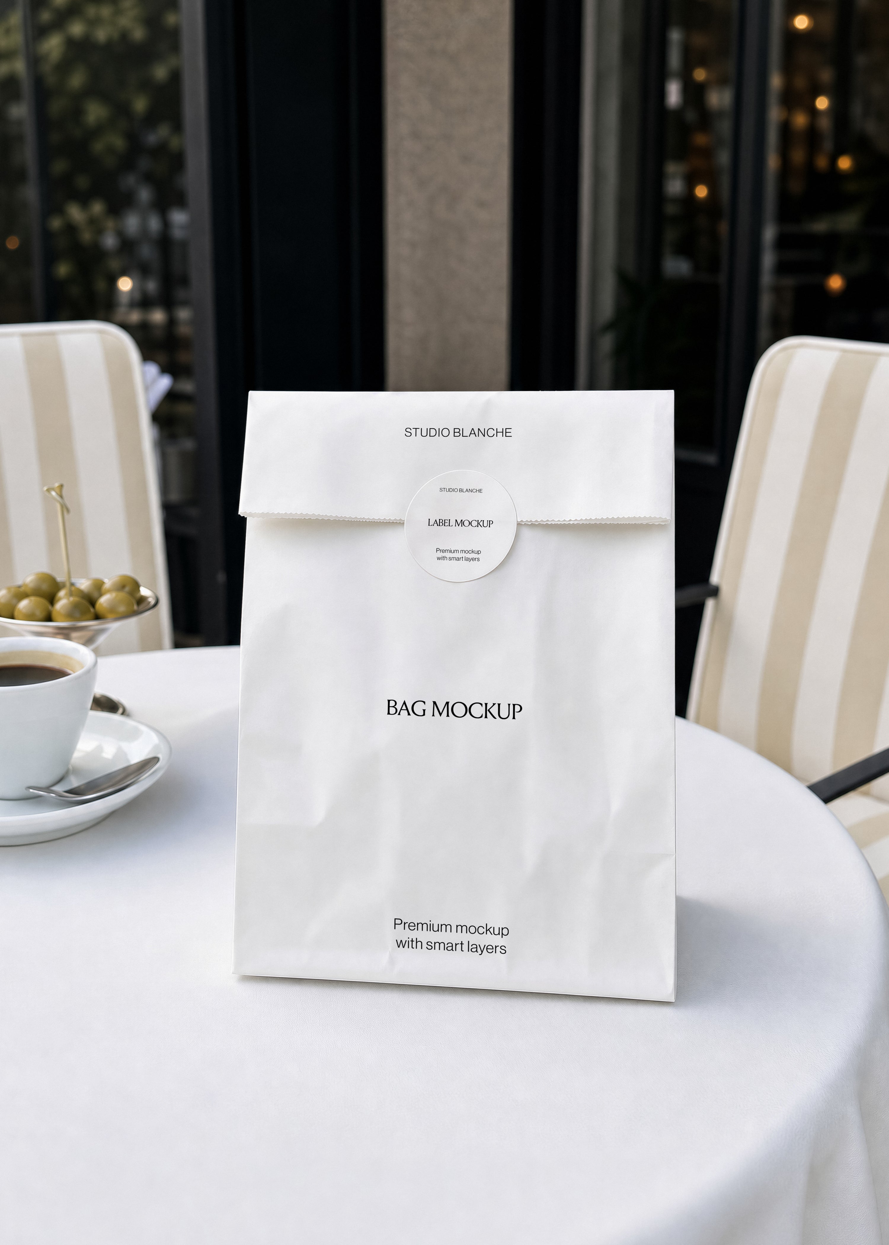 Paper bag mockup with editable label and text areas on white tablecloth, cream striped chairs and dark windows behind