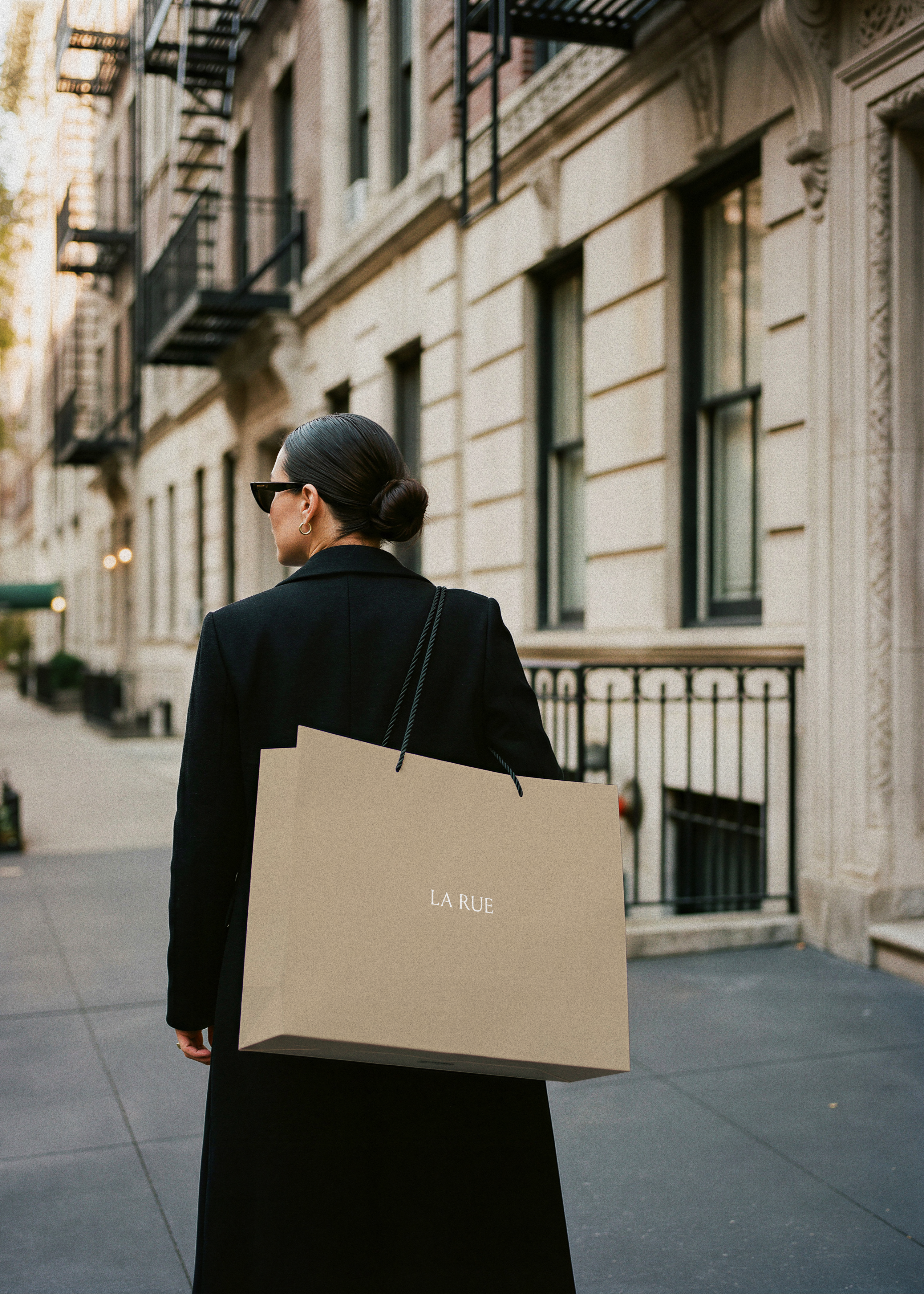 Woman in black coat carrying a tan luxury shopping bag with serif brand name on a New York City street, from Studio Blanche