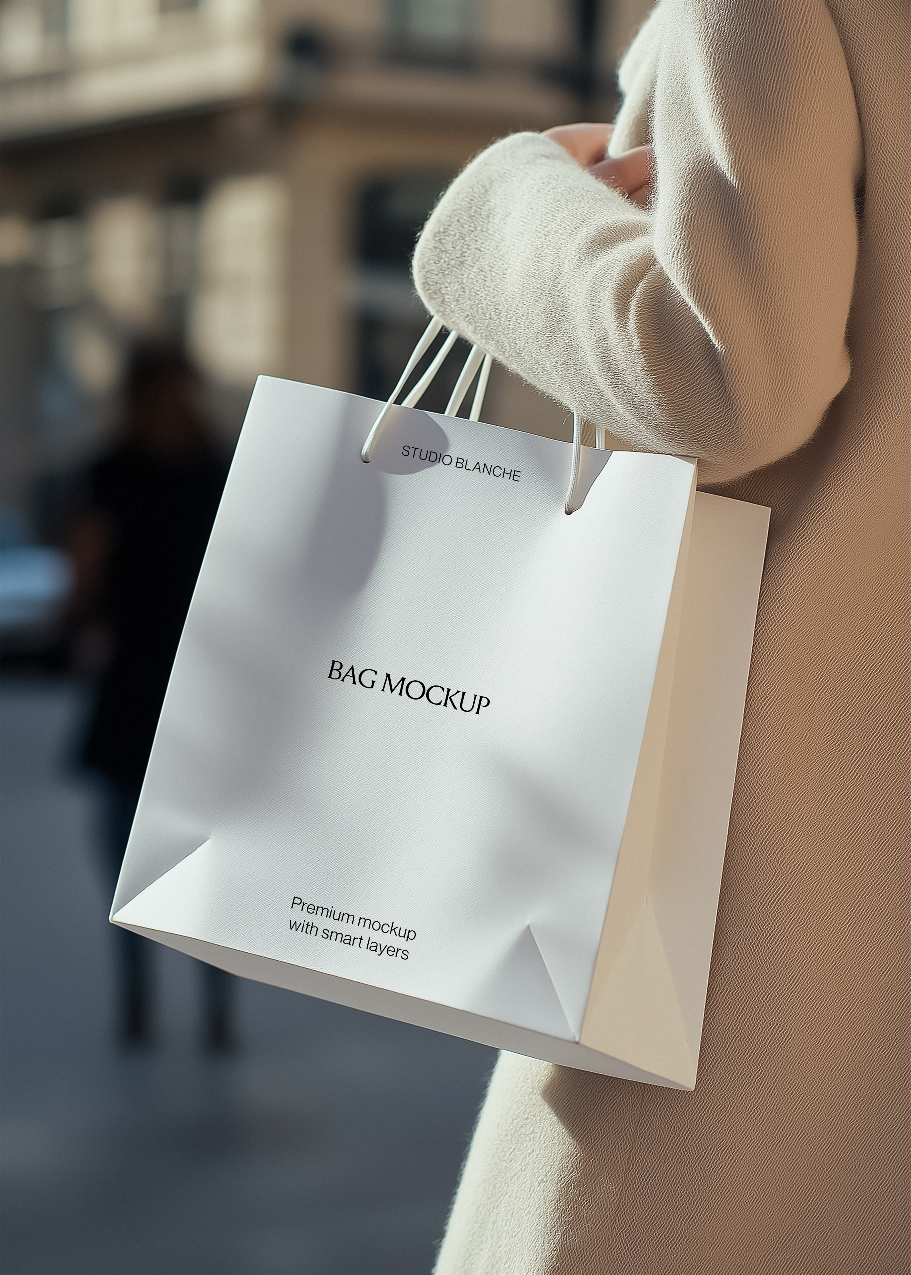 White paper shopping bag mockup with rope handles held against a cream wool coat, sunlit blurred street behind