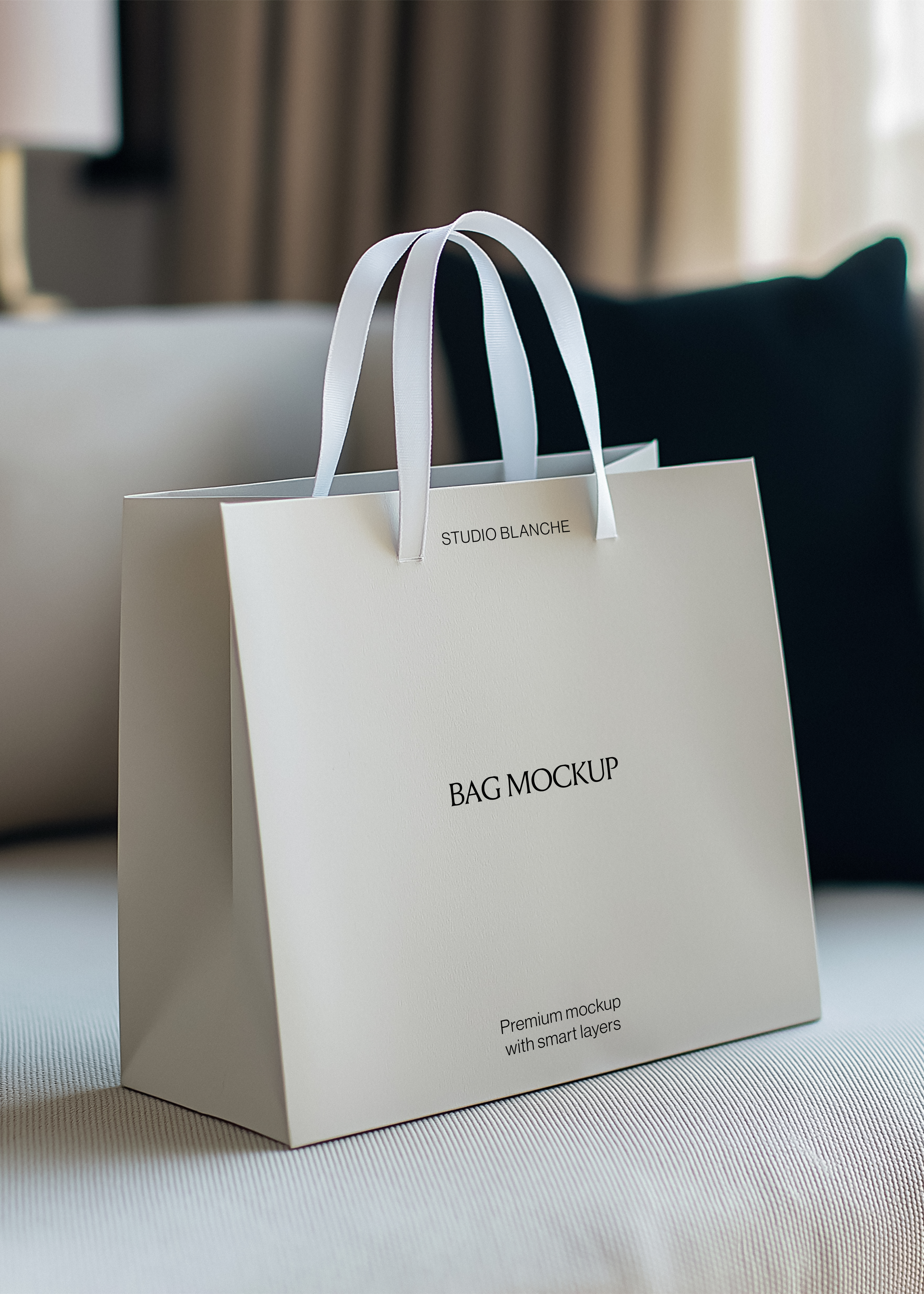 Cream paper shopping bag mockup with satin handles on a pale sofa, black cushion, warm curtains and lamp behind