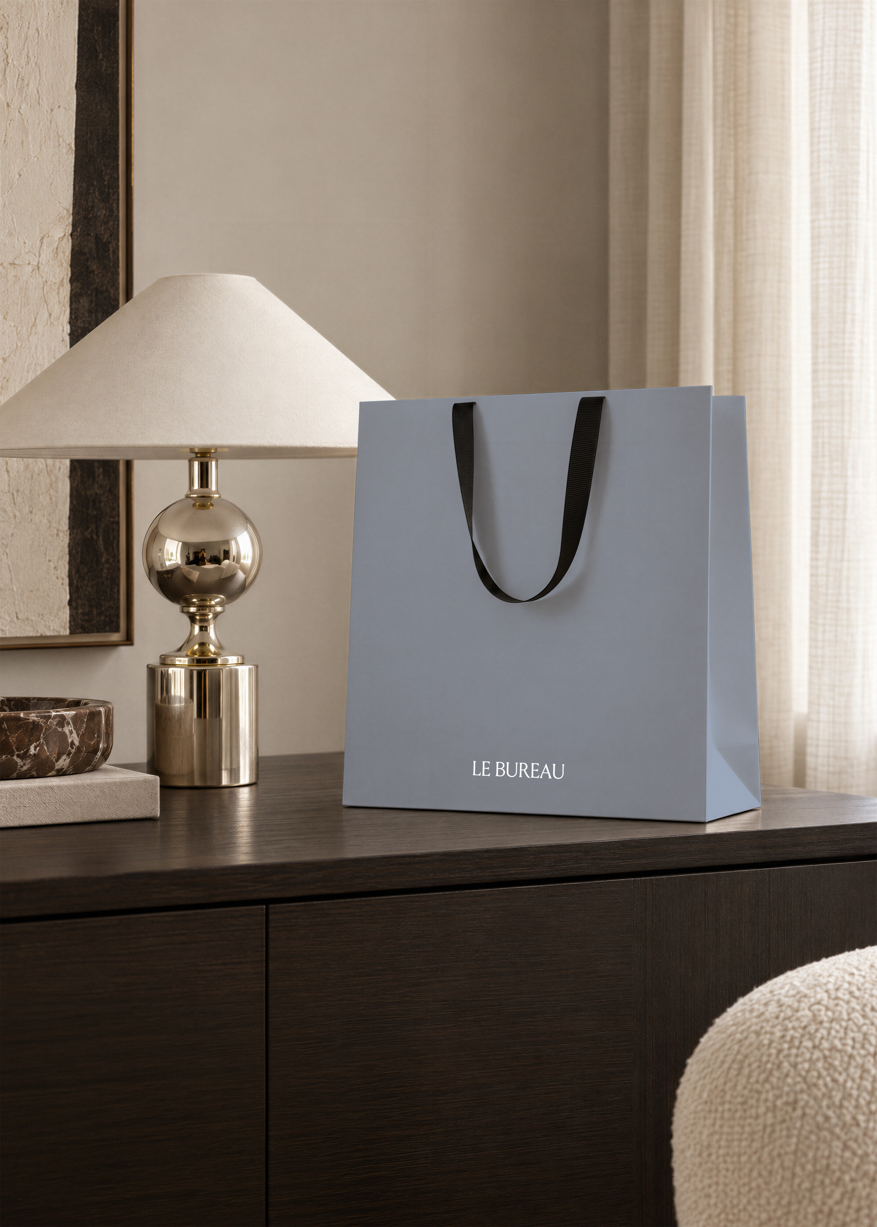 Powder blue paper shopping bag mockup with white logo, framed textured artwork and boucle pouf in warm neutral room