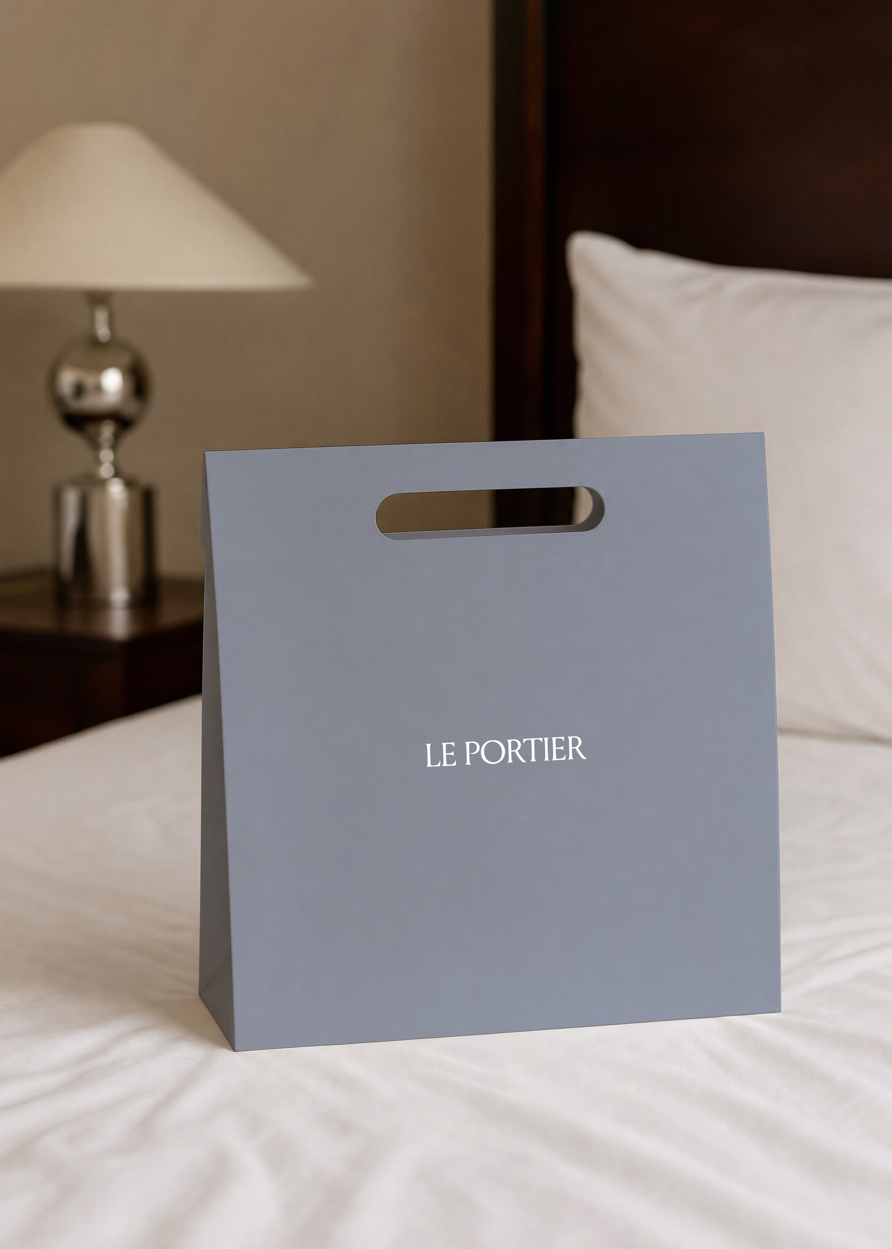 Paper shopping bag mockup standing upright on a white hotel bed with pillow, dark wood headboard behind