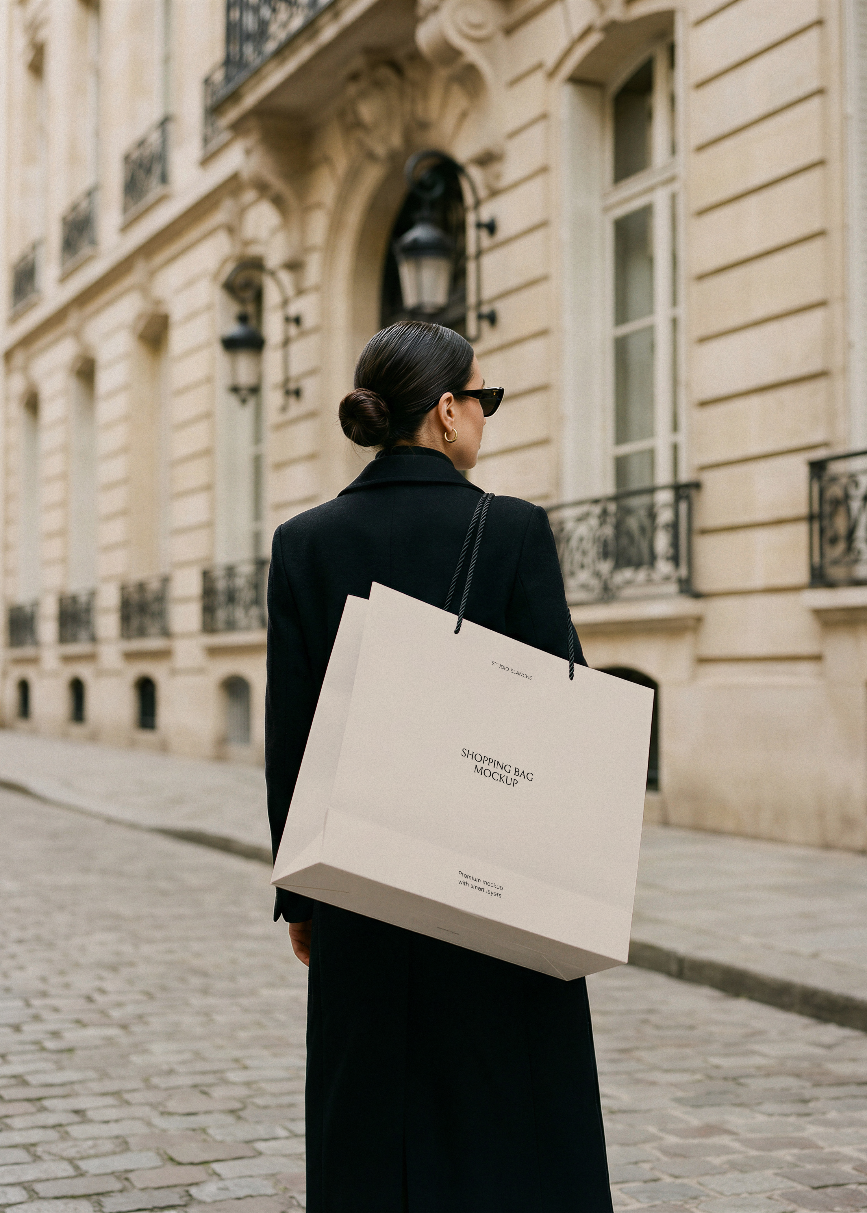 Woman in black coat carrying a blank cream luxury shopping bag with rope handles on a cobblestone Paris street, from Studio Blanche