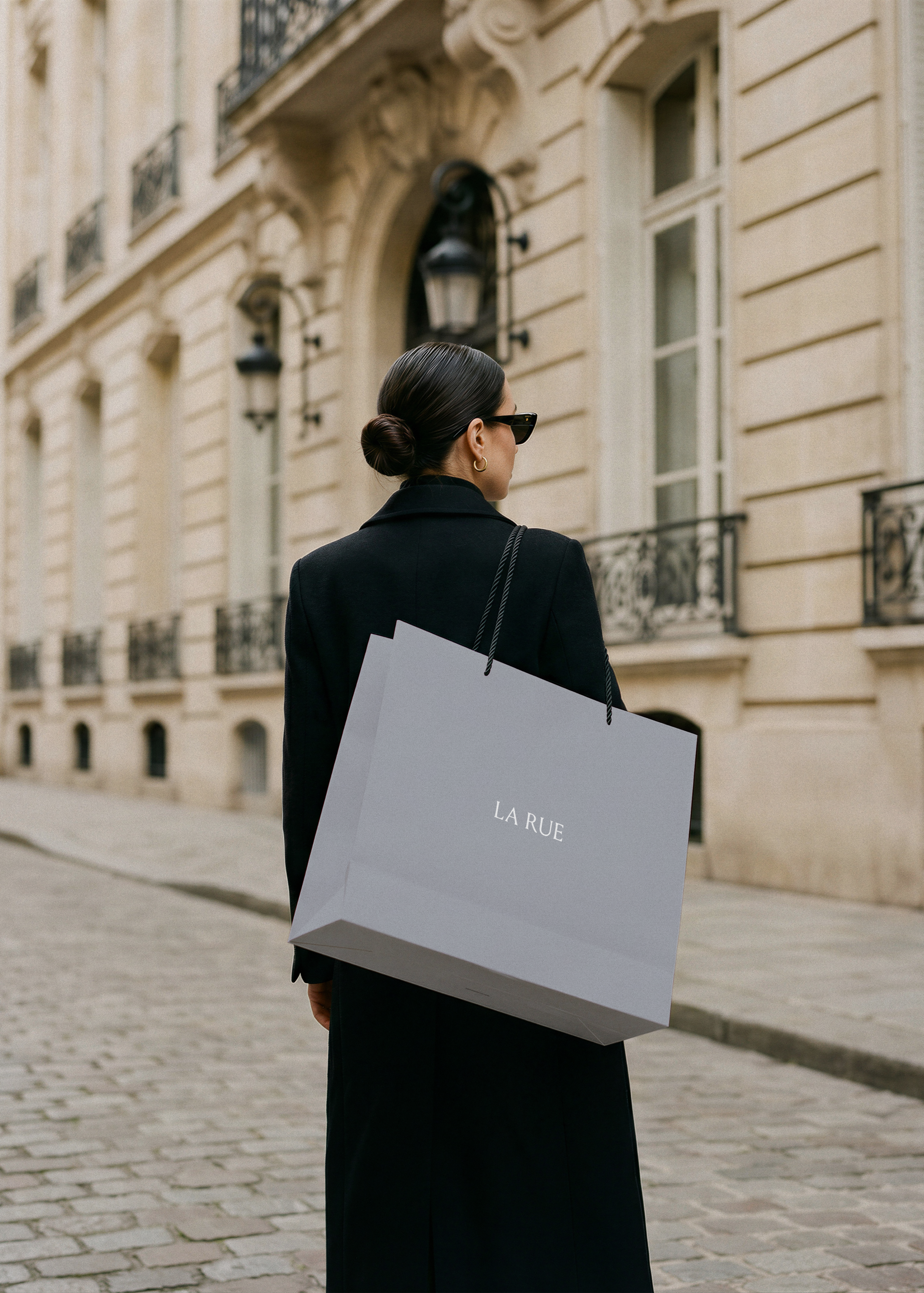 Woman in black coat carrying a light grey luxury shopping bag with serif brand name on a cobblestone Paris street, from Studio Blanche