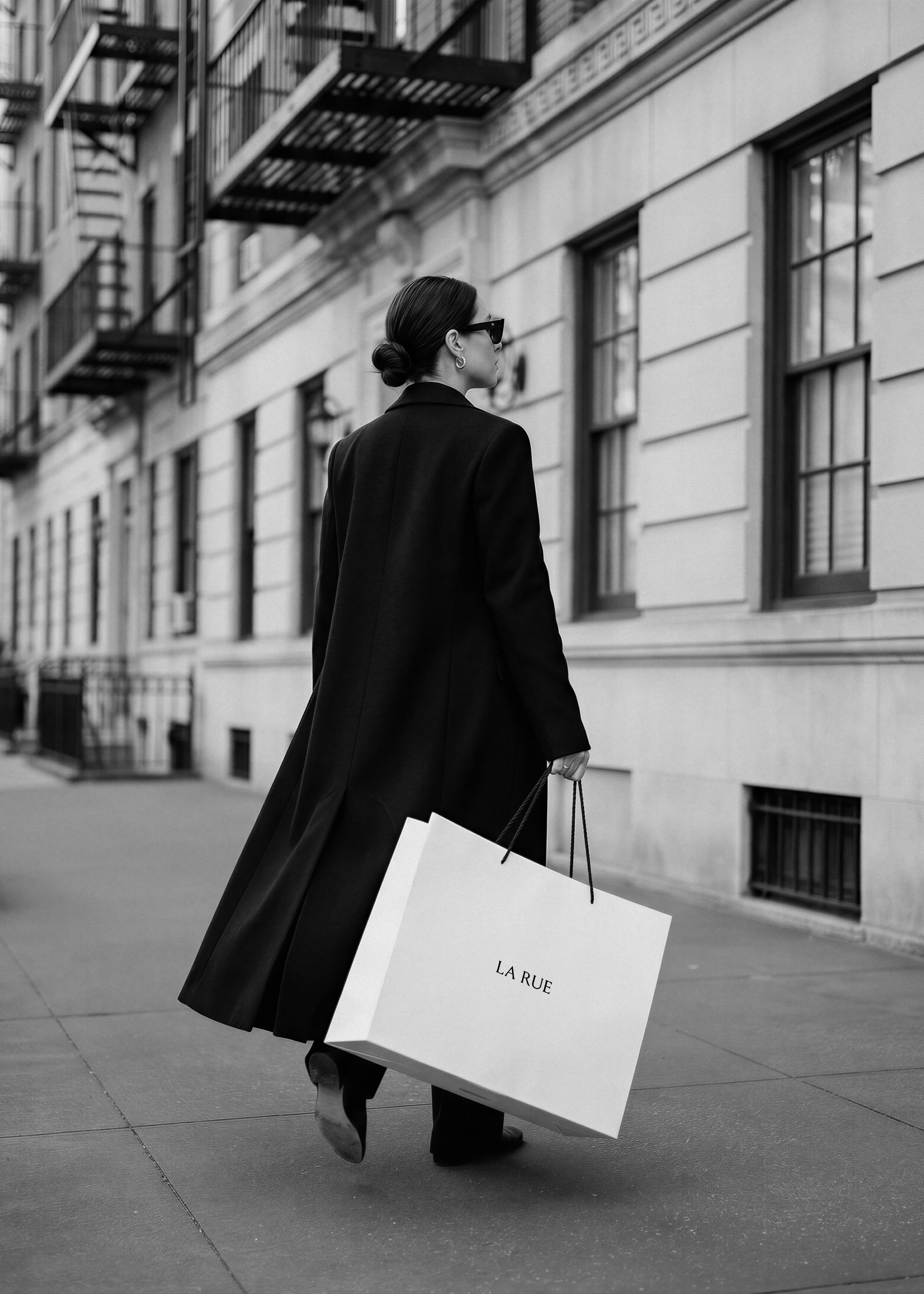 Paper shopping bag with black logo and twisted handles held mid-stride against a monochrome brownstone street backdrop