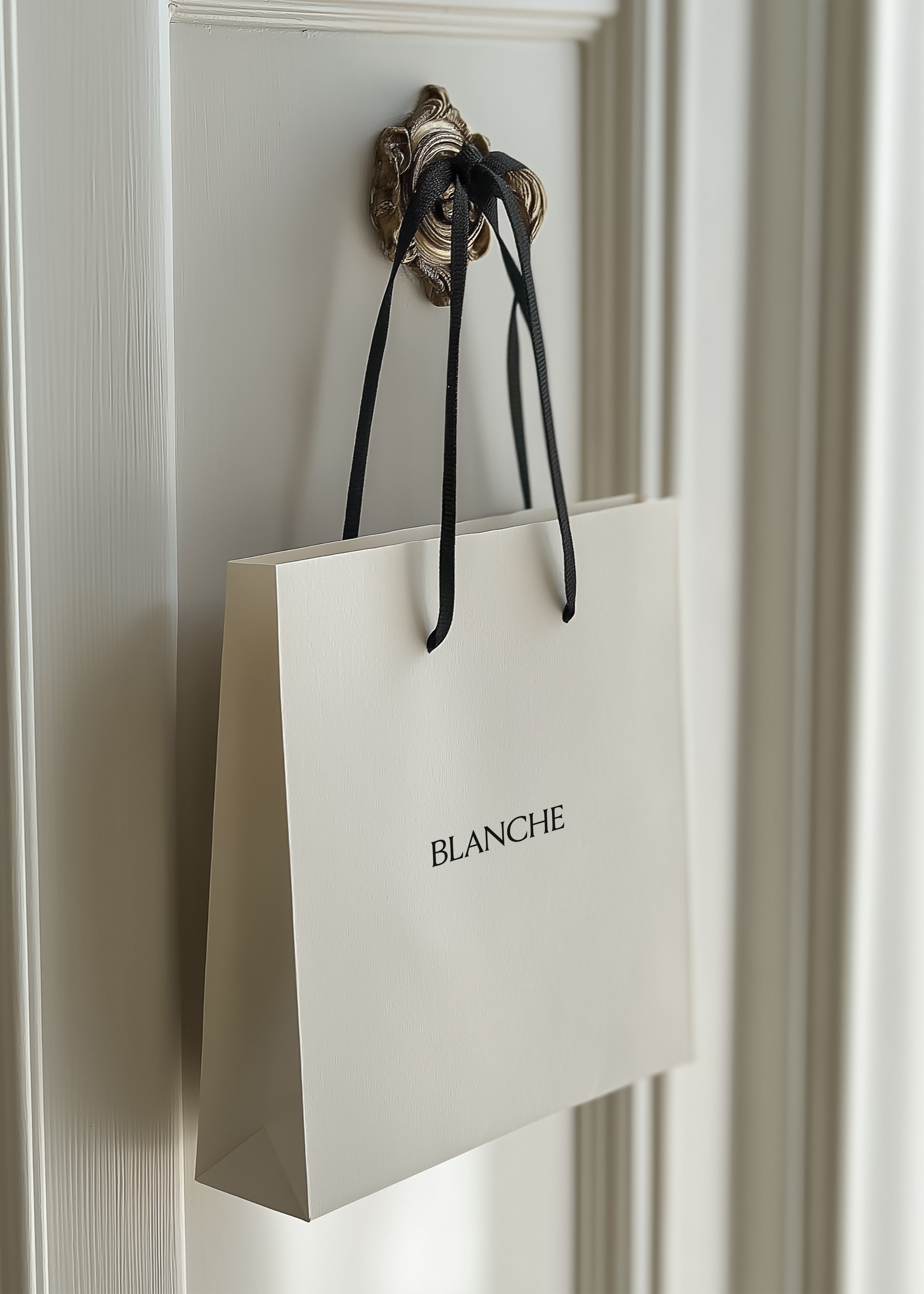 Cream paper shopping bag with black ribbon handles and Blanche logo hung from a brass handle on a white panelled door