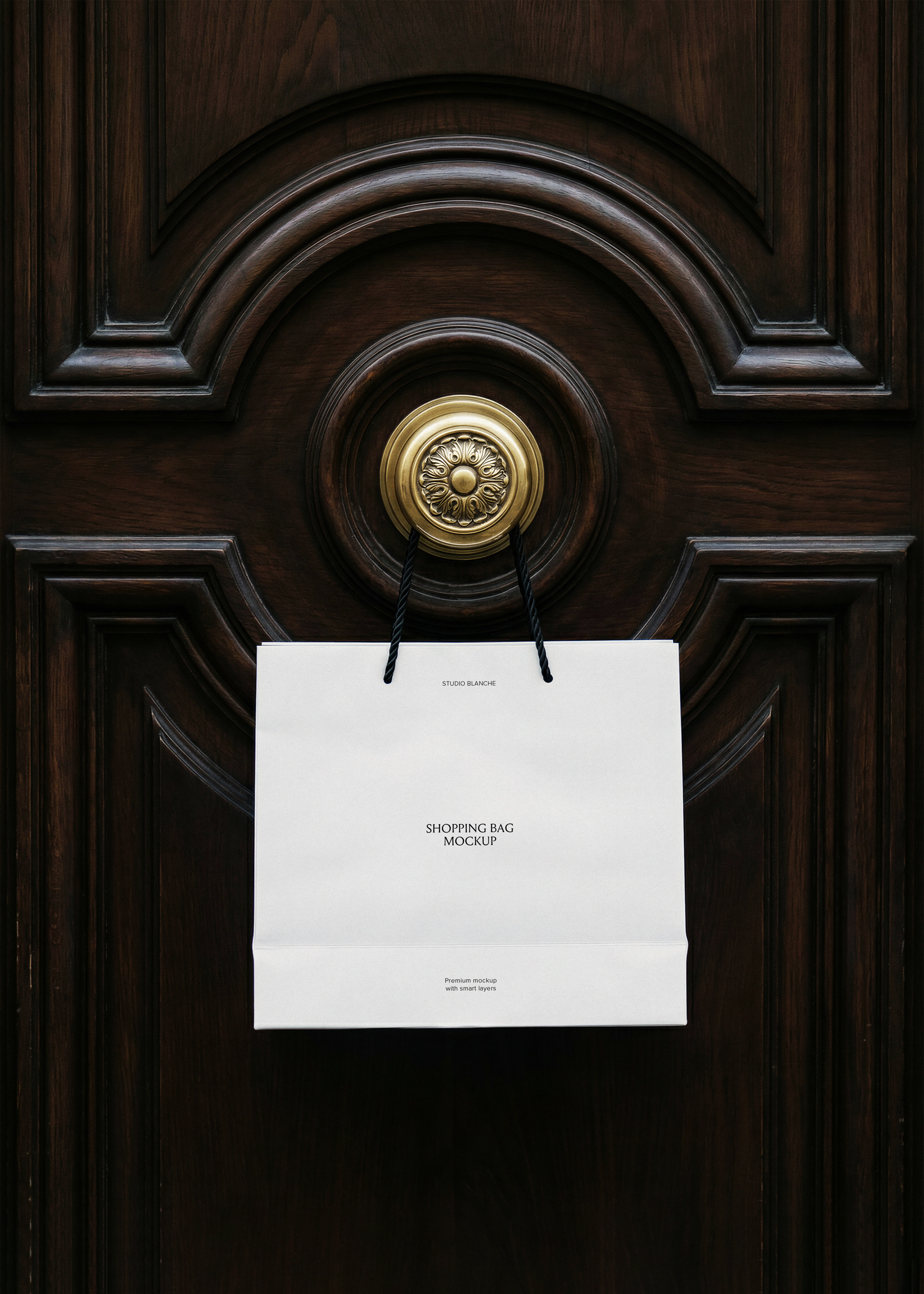 Blank white luxury shopping bag with black rope handles hanging from an ornate brass door knob on dark carved mahogany panelling, from Studio Blanche