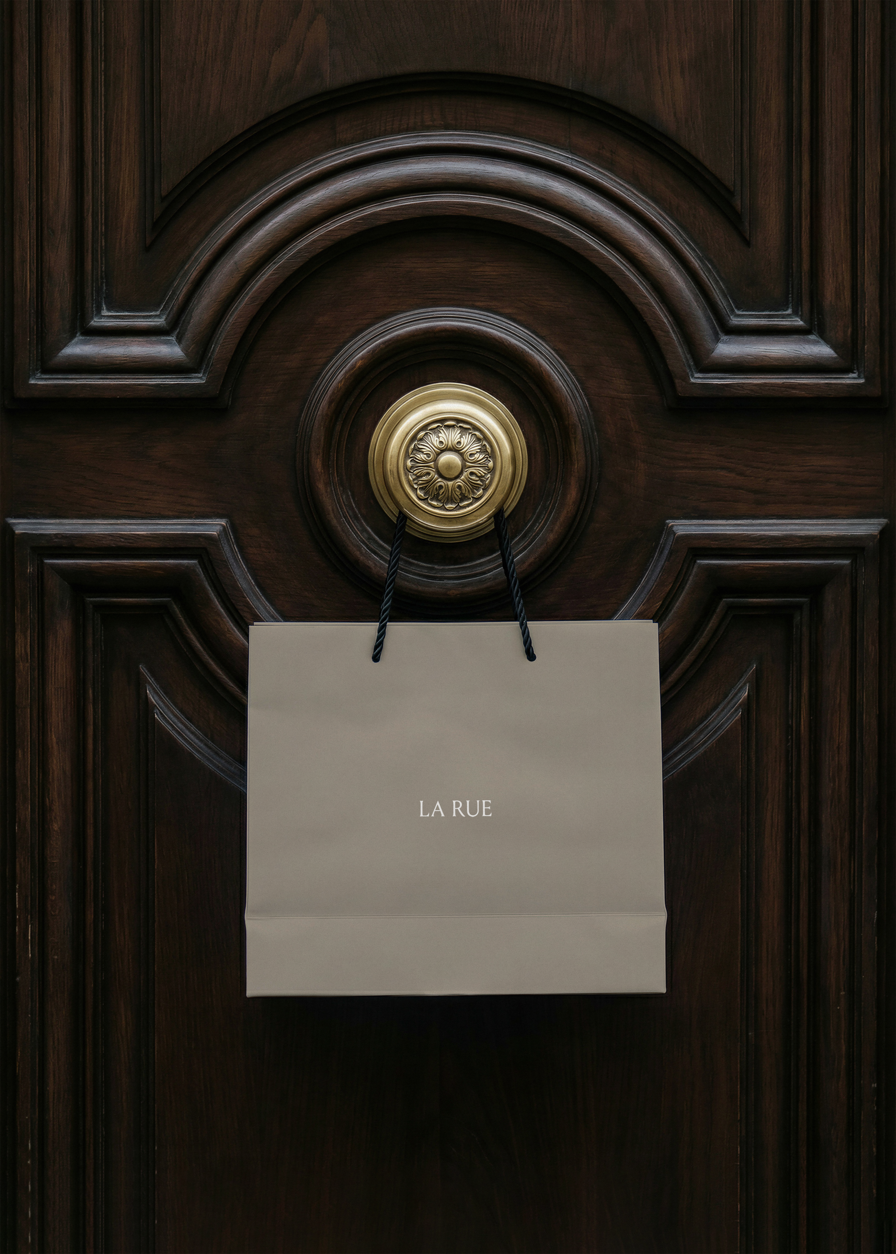 Taupe luxury shopping bag with white serif brand name hanging from an ornate brass door knob on dark carved mahogany panelling, from Studio Blanche