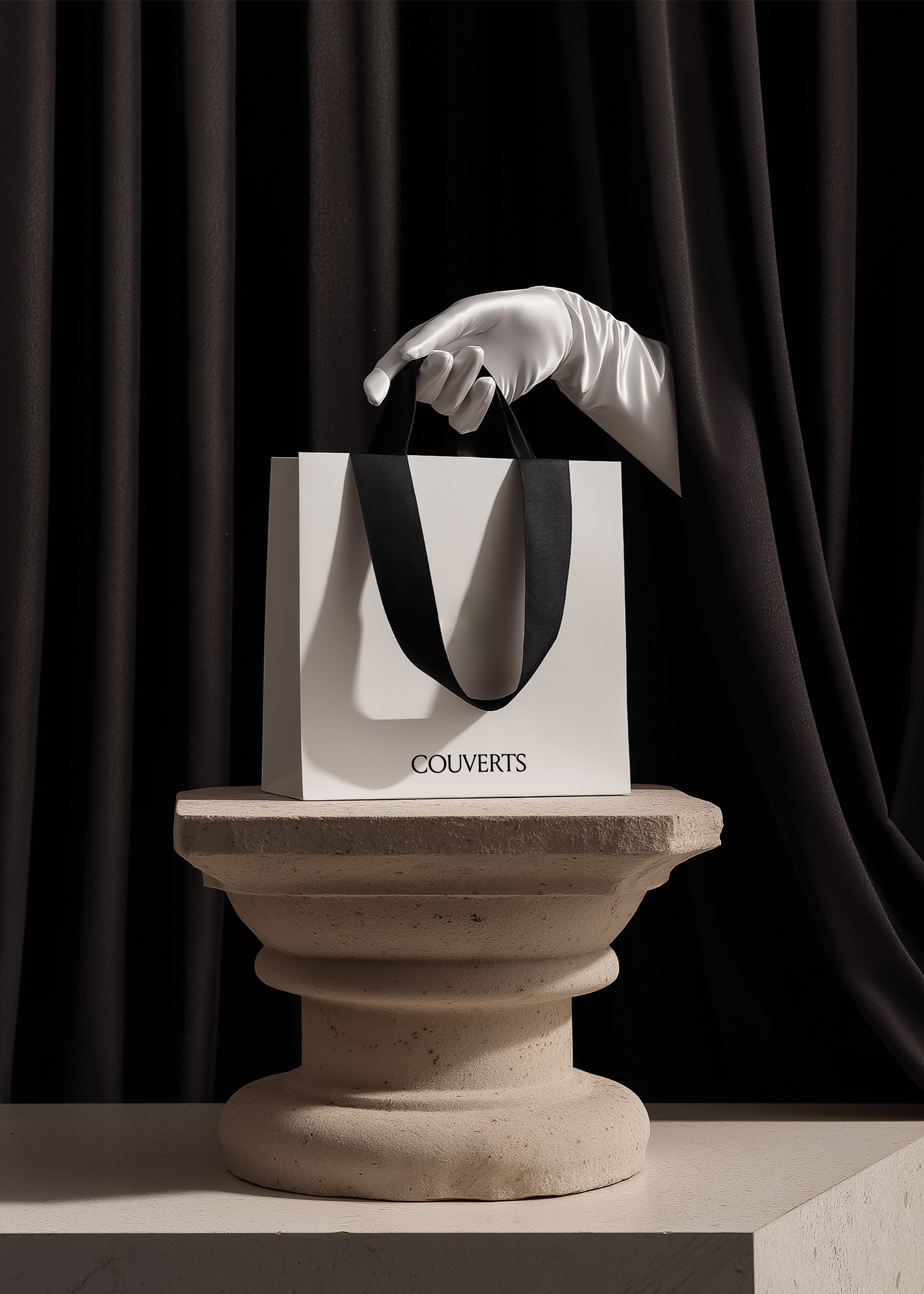 White paper shopping bag with black ribbon handles held by white satin-gloved hand on rough stone column pedestal, dark draped curtain background