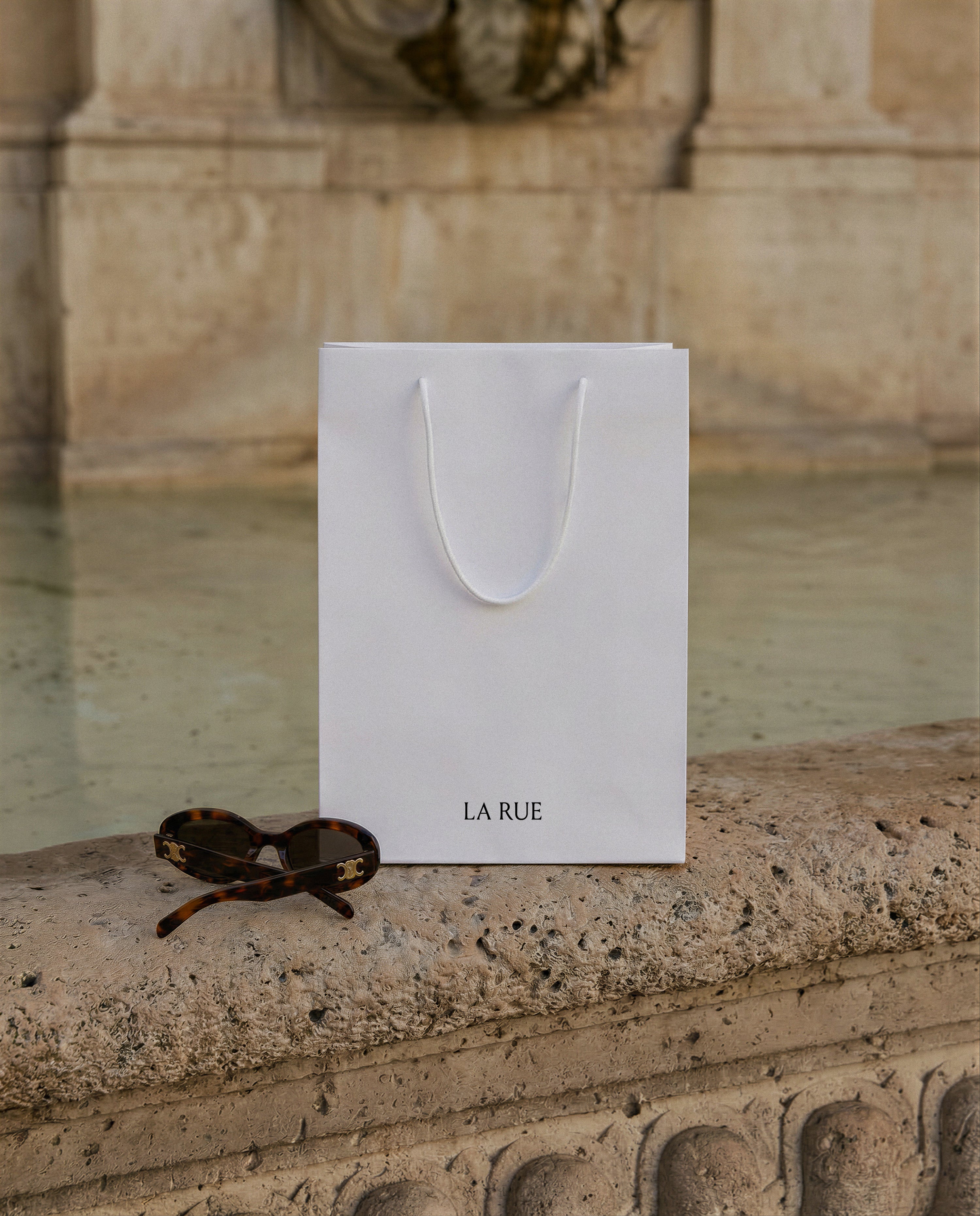 White luxury shopping bag with serif brand name on a carved stone fountain ledge beside tortoiseshell sunglasses, from Studio Blanche