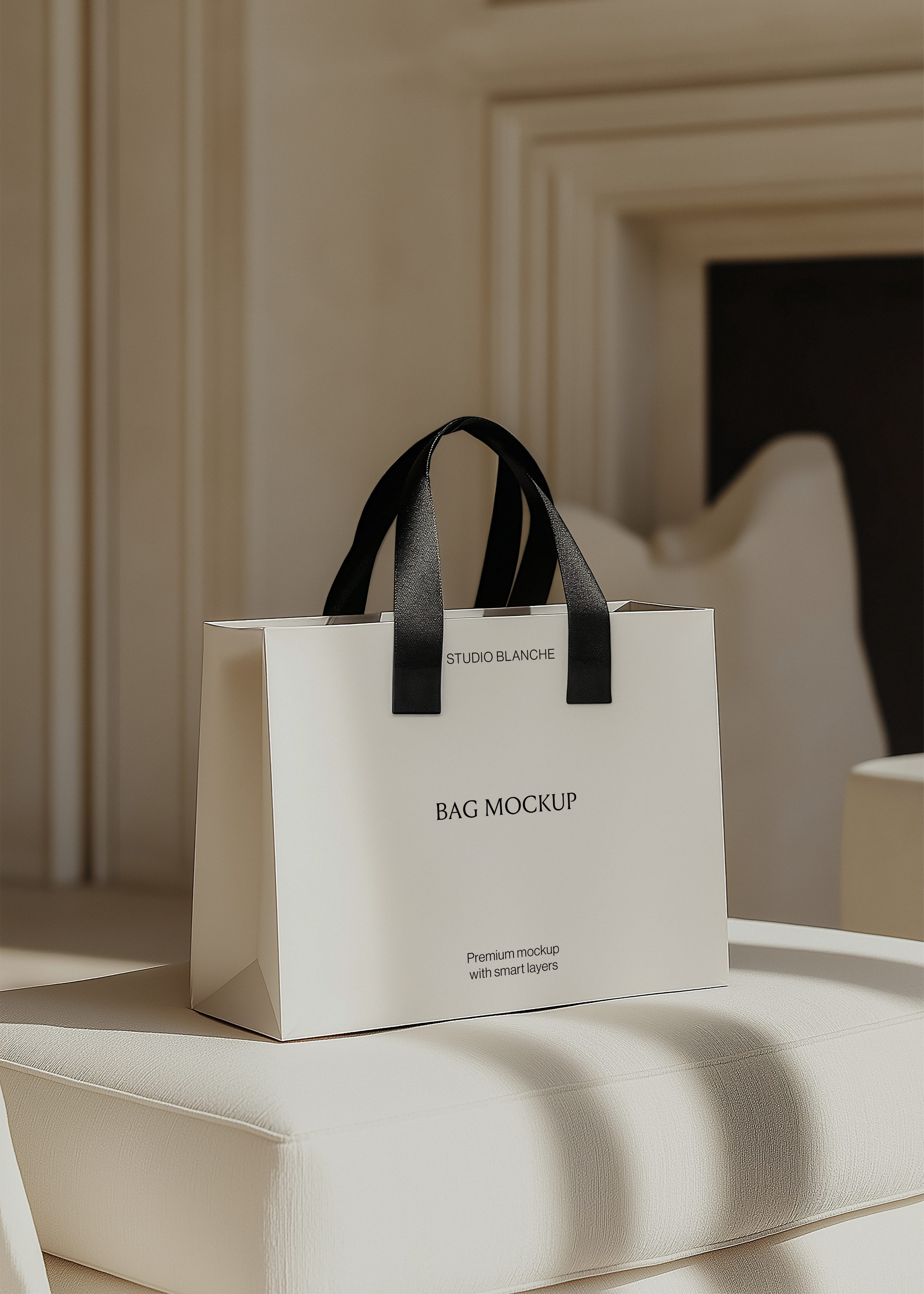 White paper shopping bag mockup with black satin handles on a cream ottoman, moulded interior wall and framed art behind