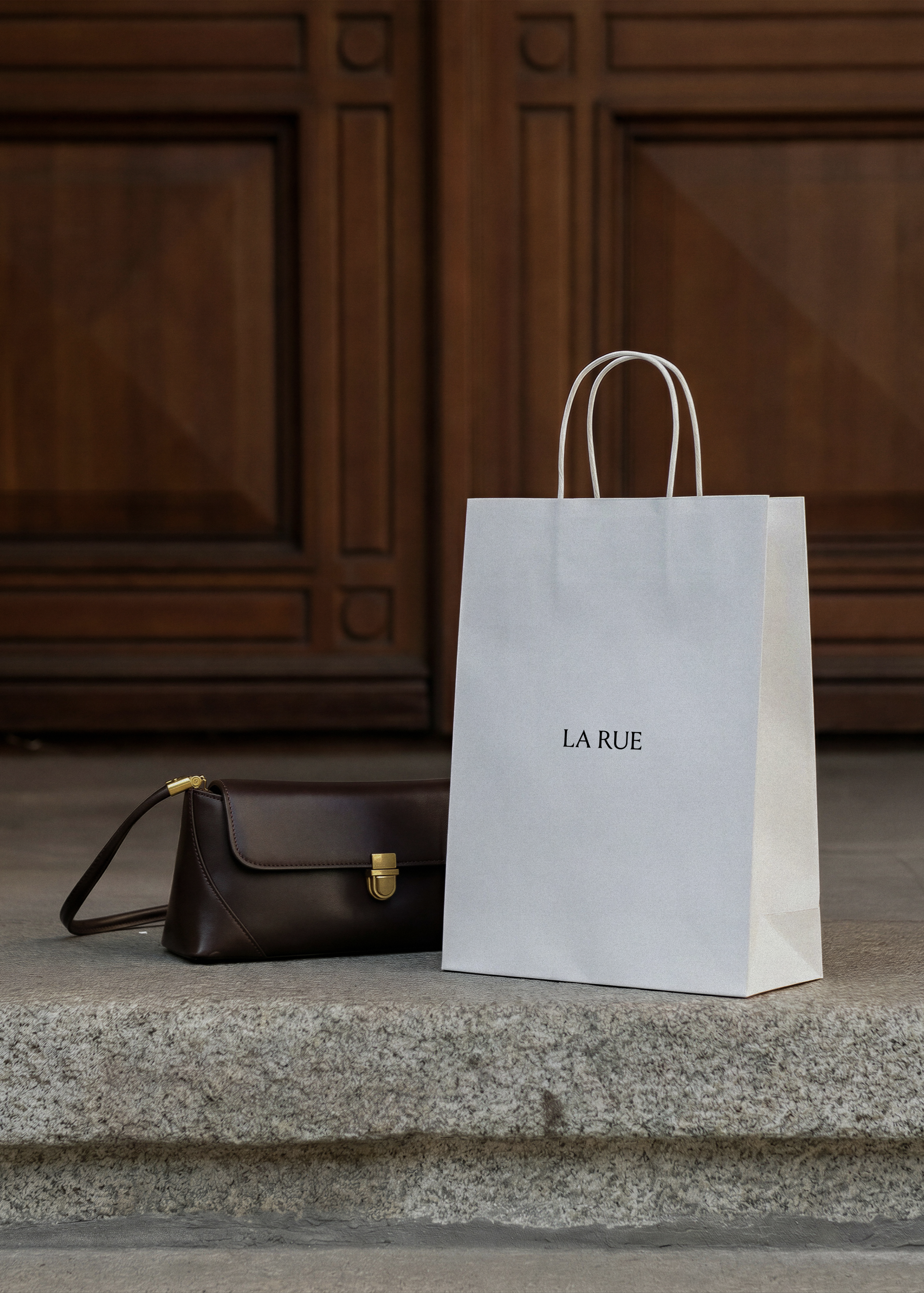 Shopping bag mockup standing on stone step beside dark brown leather handbag with gold clasp against carved wooden door