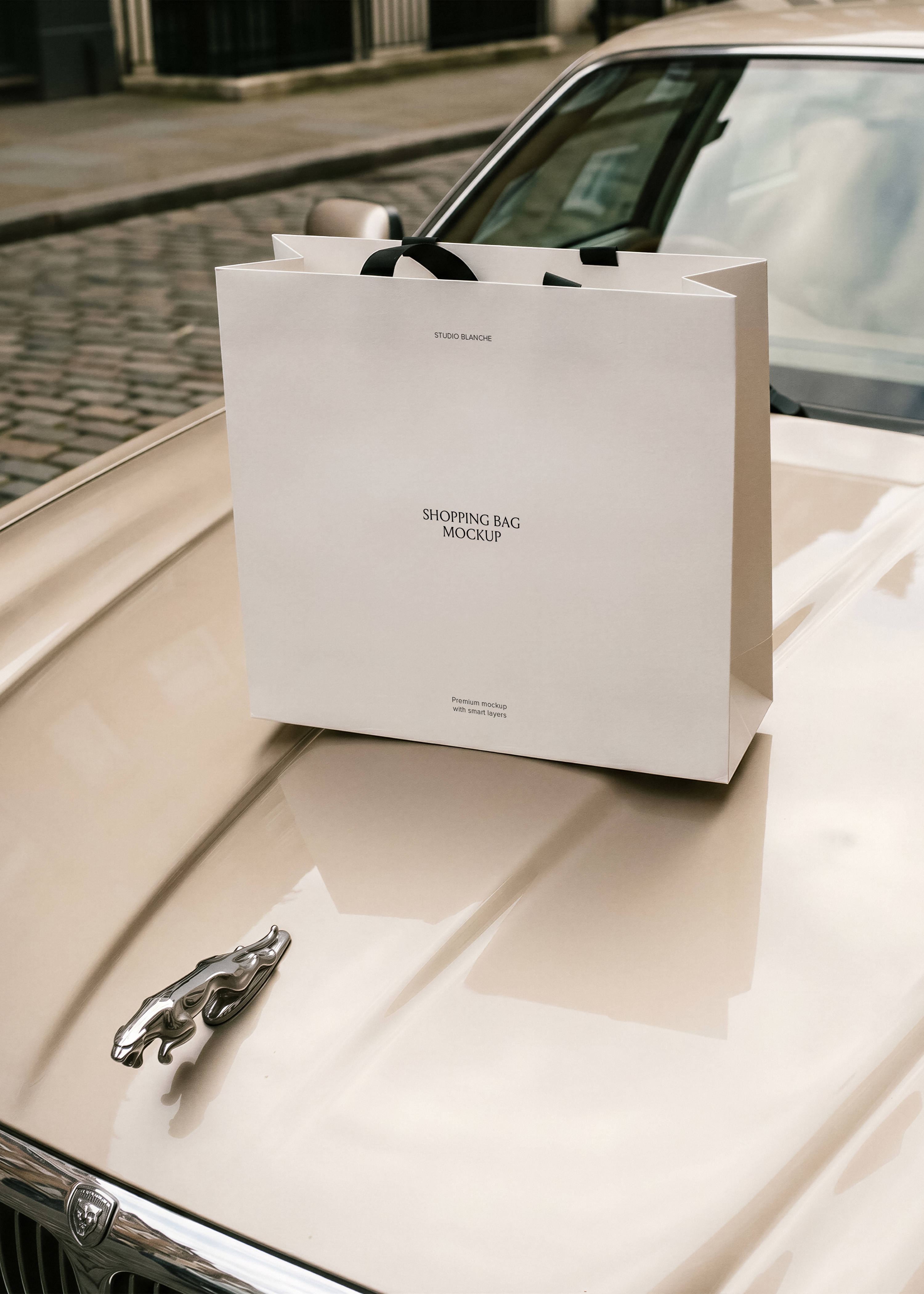 White paper shopping bag mockup with black ribbon handles styled on glossy cream car bonnet beside silver leaper emblem