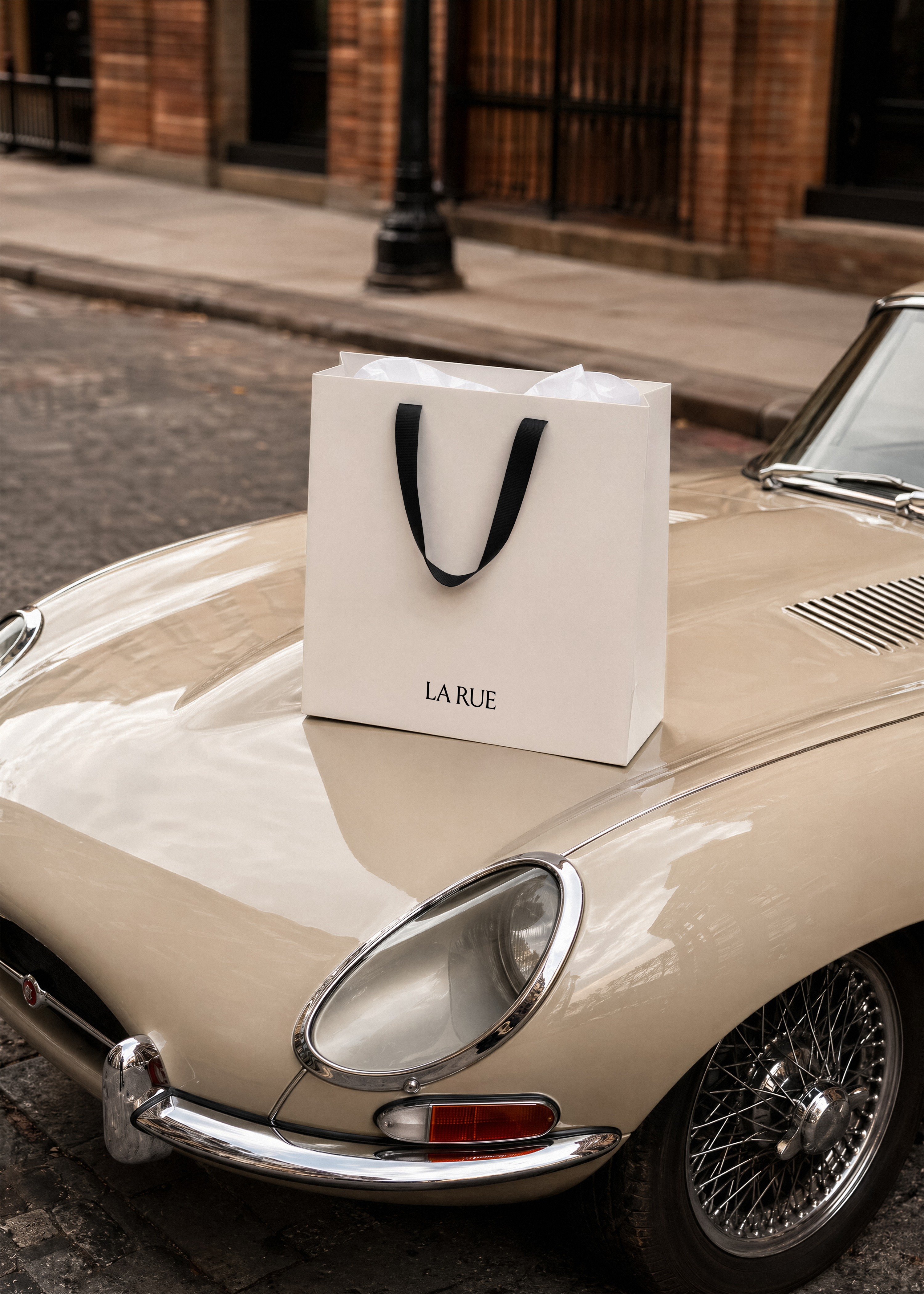 White LA RUE shopping bag with black ribbon handles on the cream bonnet of a vintage Jaguar on a cobbled street