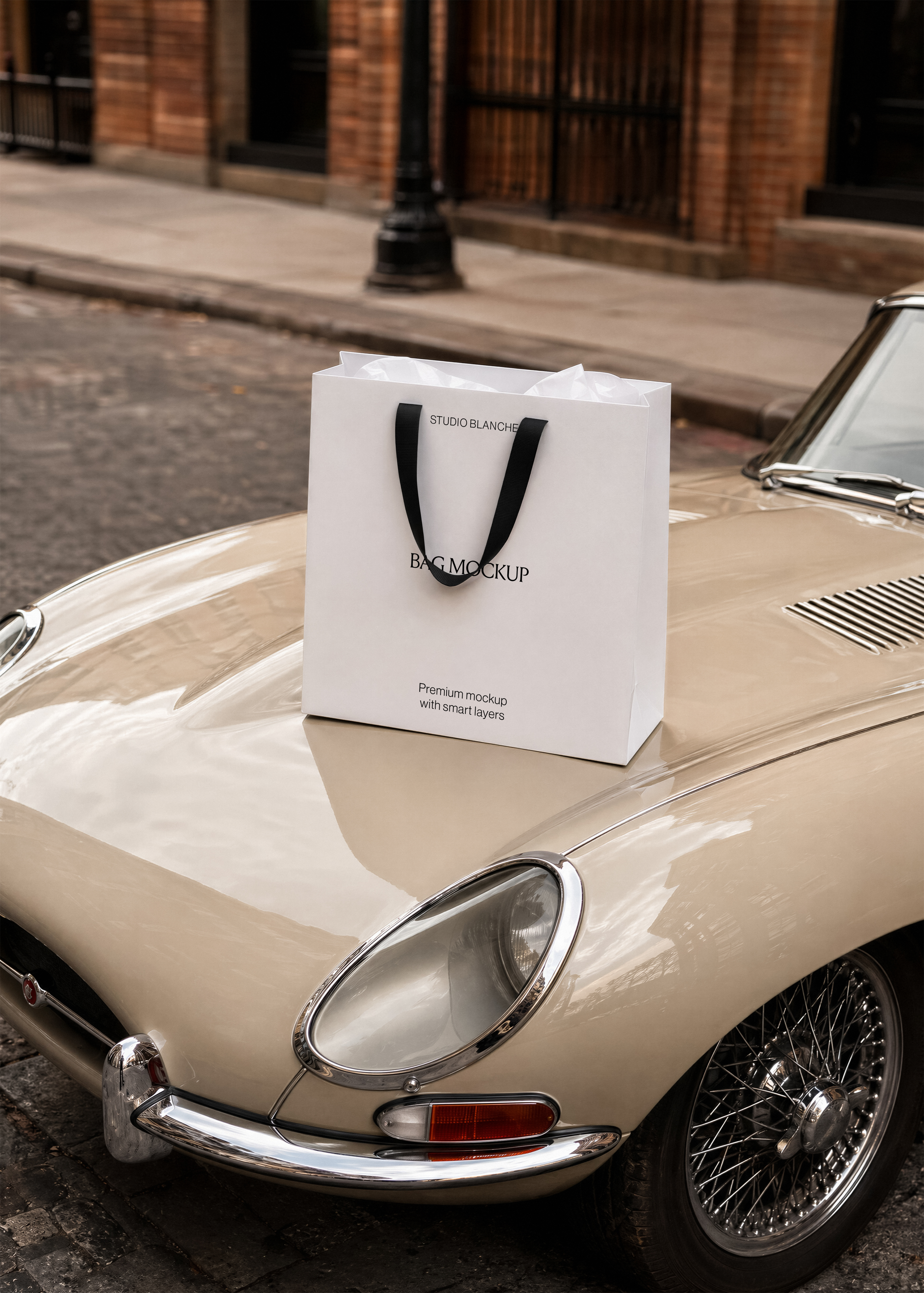 Studio Blanche paper bag mockup with black handles standing on a glossy cream vintage car bonnet, brick building behind