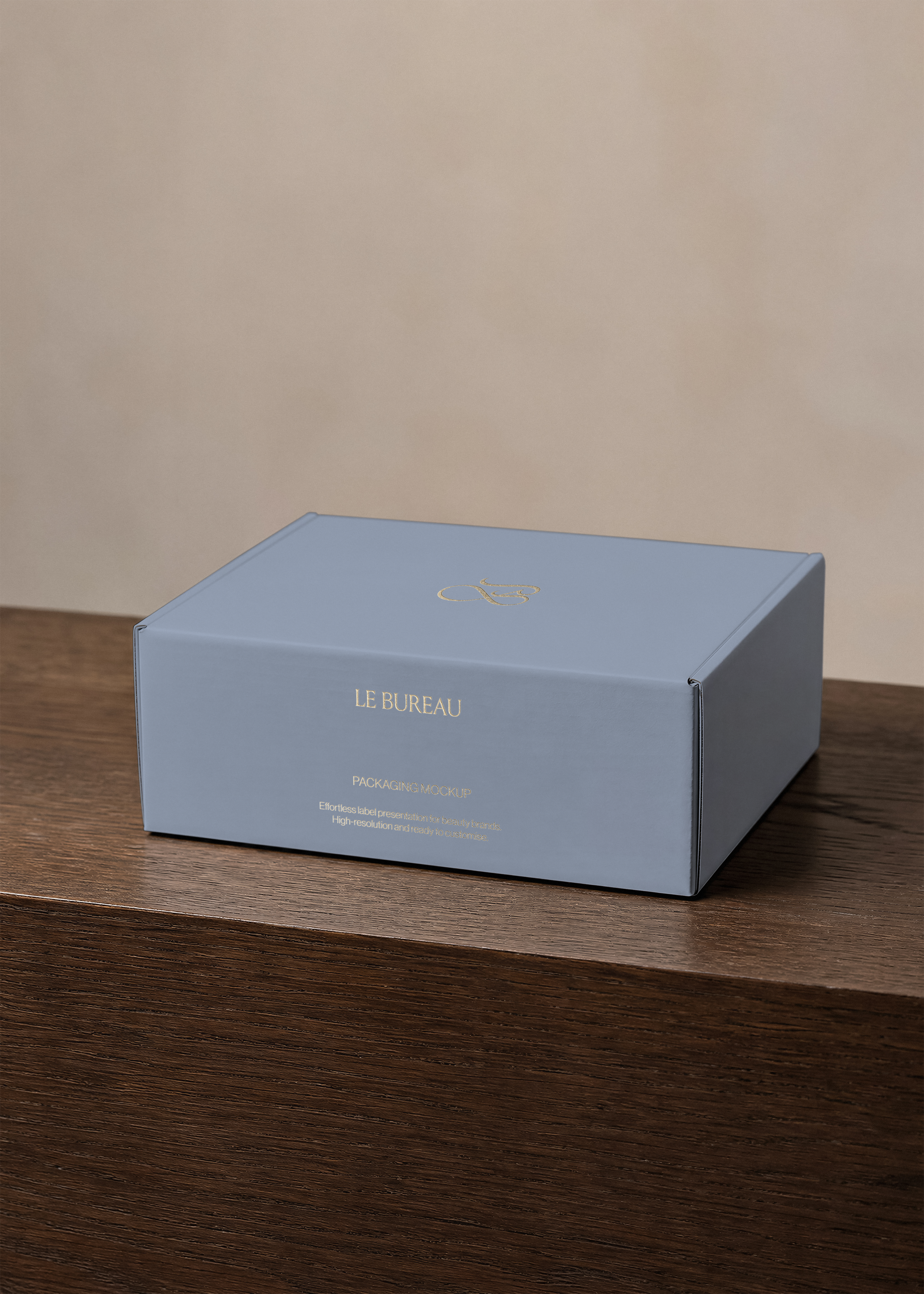 Dusty blue packaging box mockup with gold foil wordmark and monogram, angled on a dark wood surface against a warm backdrop