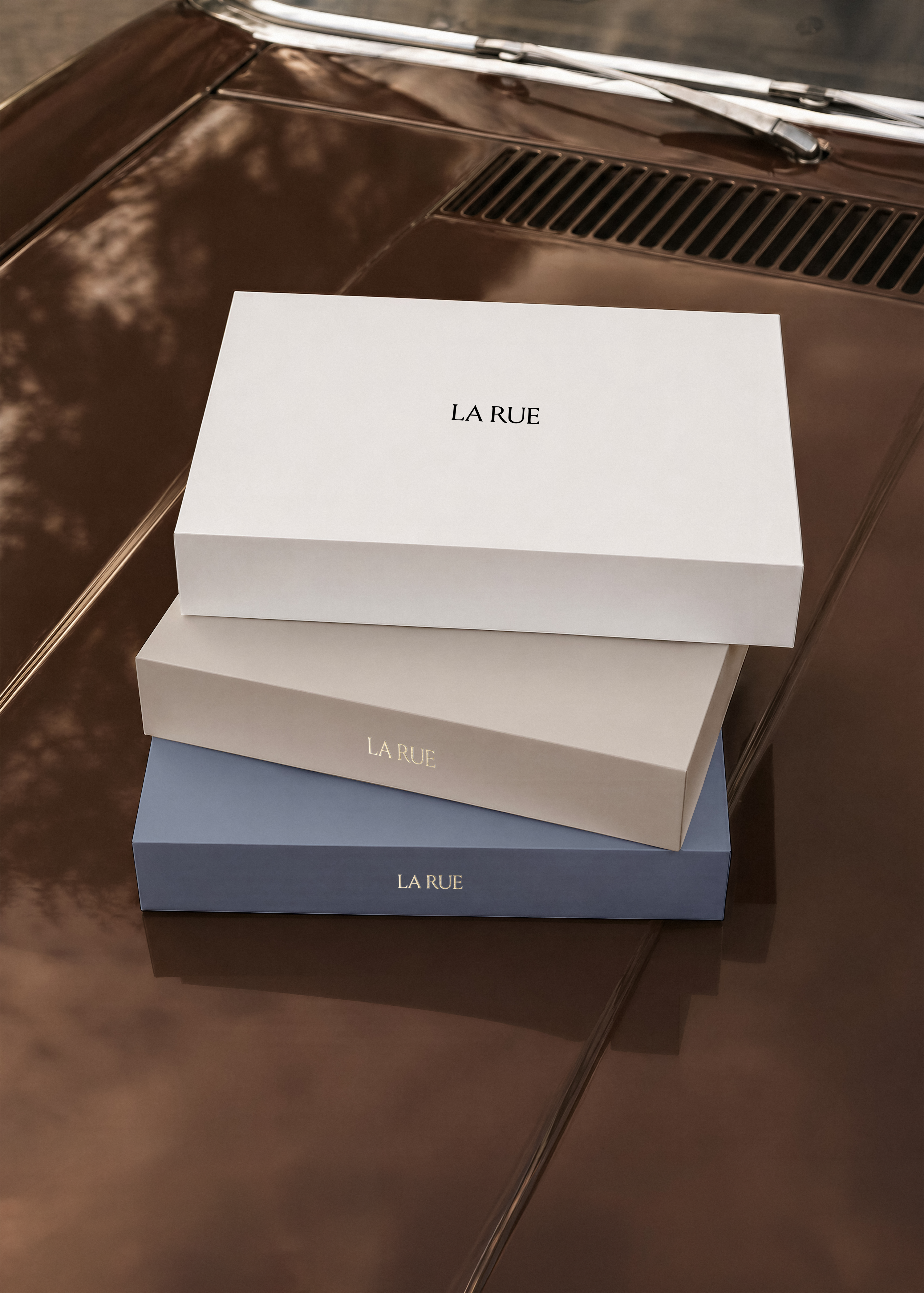 Gift box mockup, three stacked lidded boxes in white, beige and blue on a glossy chocolate brown car bonnet