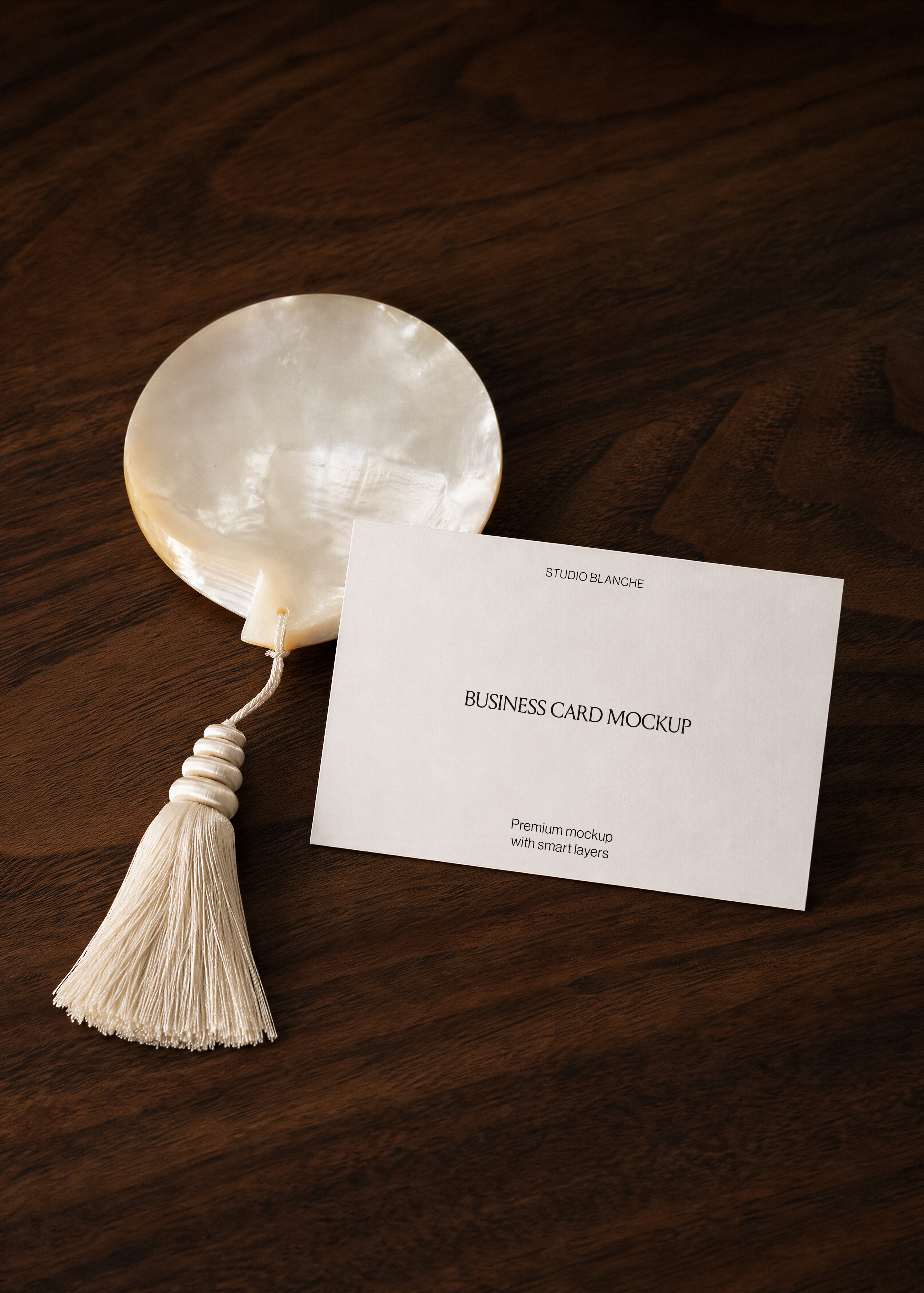 Business card mockup flat on dark walnut surface beside iridescent mother-of-pearl disc with cream silk tassel