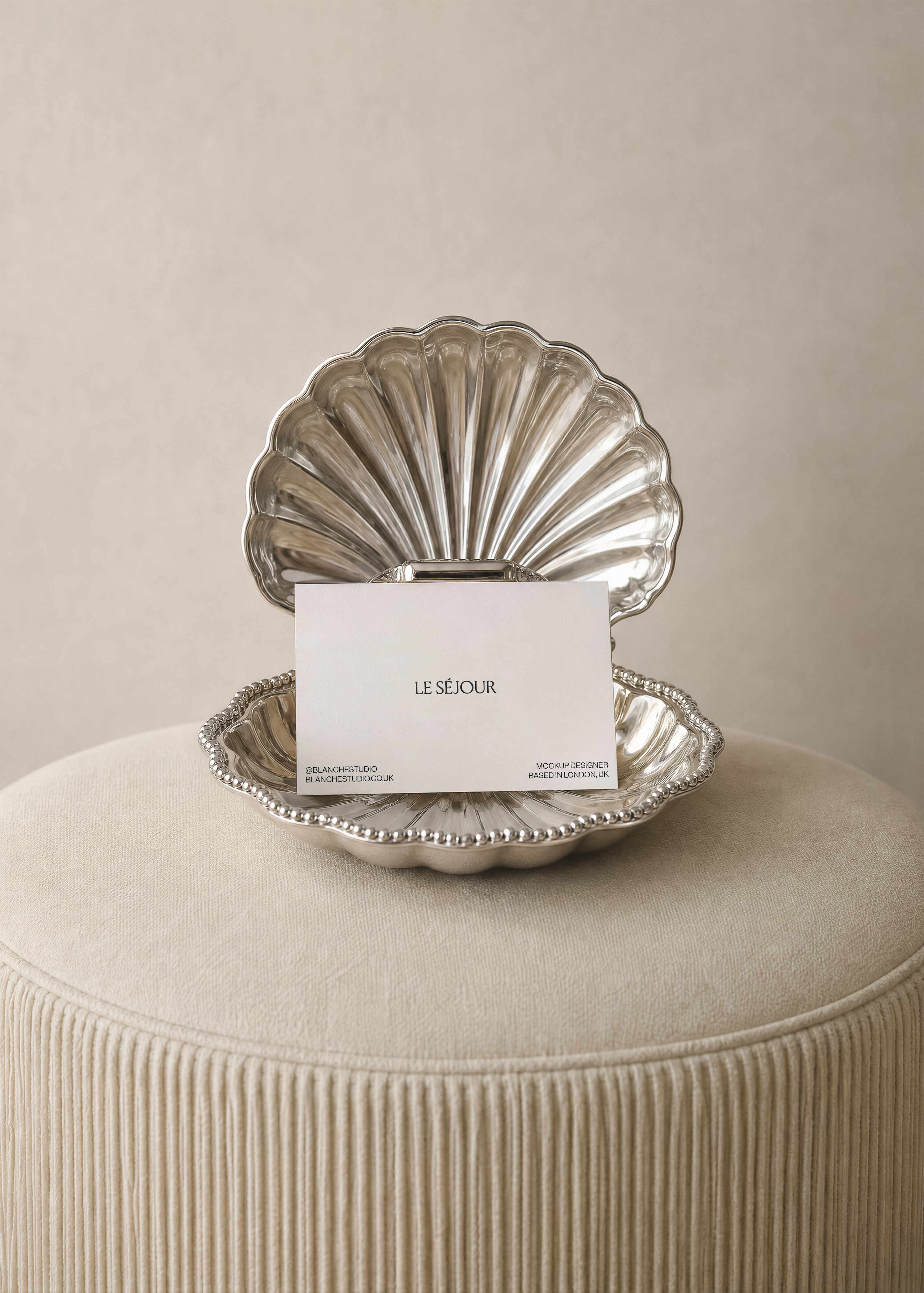 Business card mockup propped in an open silver scallop shell dish on a cream fringed velvet pouf against a linen wall