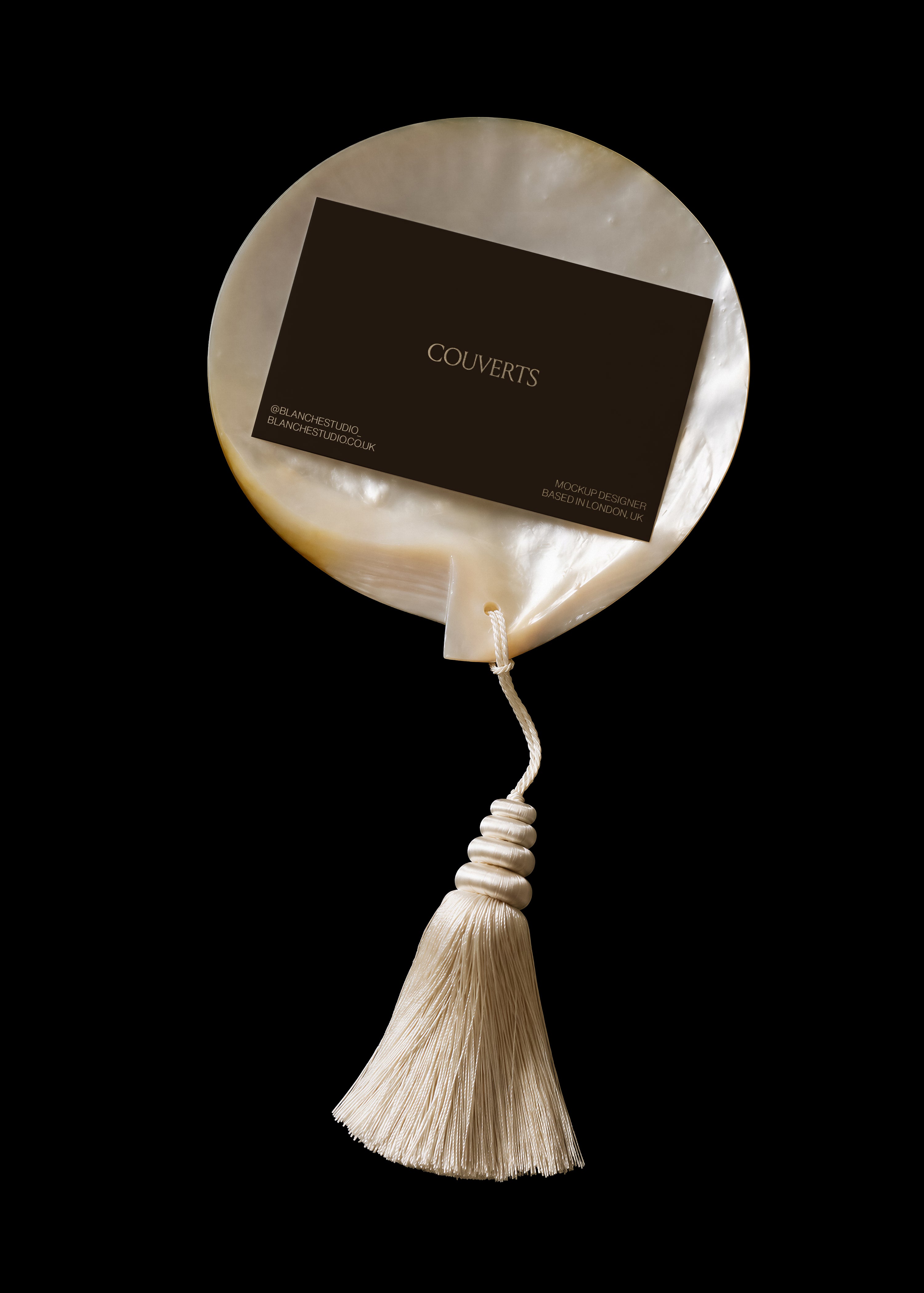 Dark business card mockup resting on a round mother-of-pearl shell with cream silk tassel below on black backdrop

