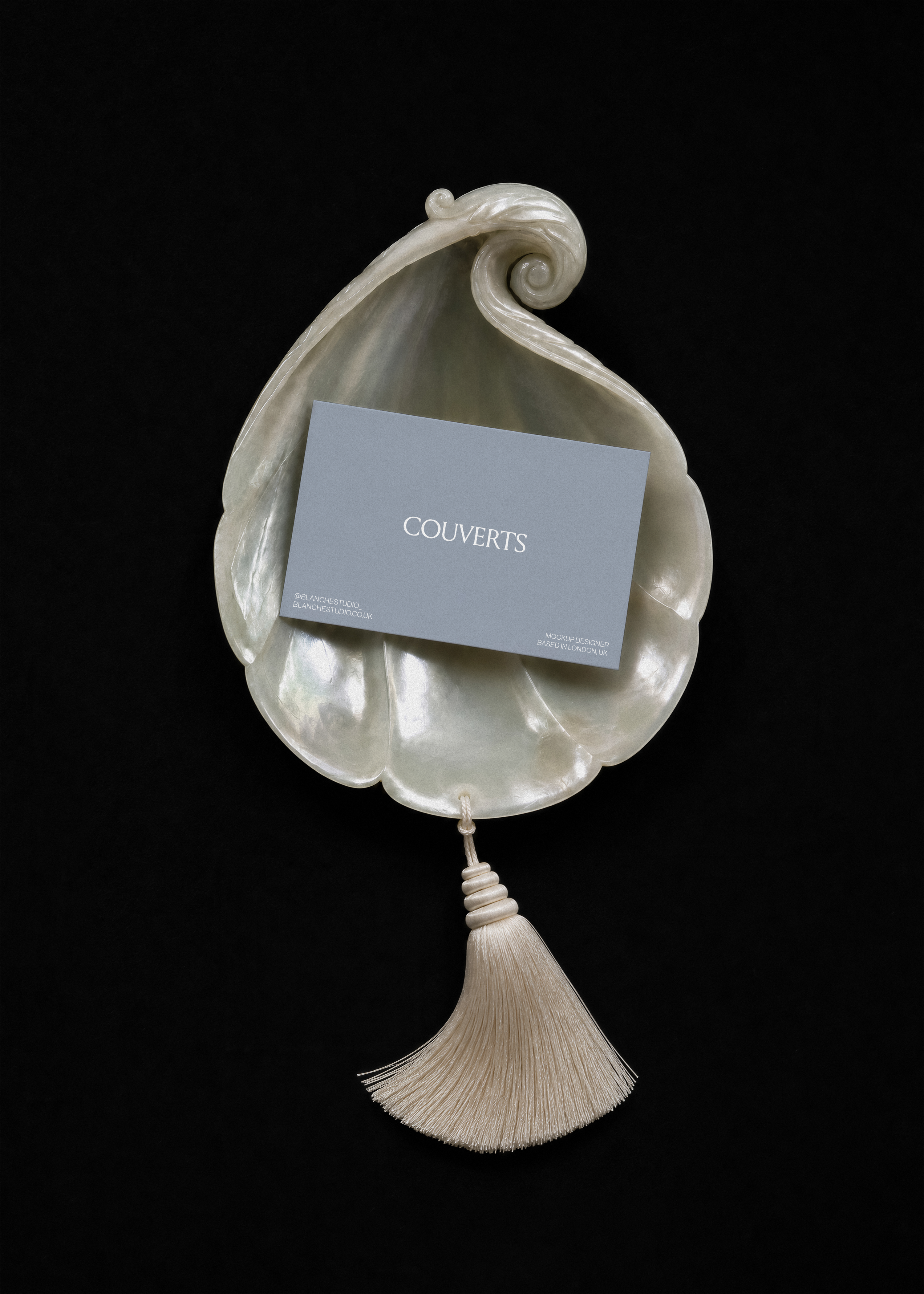 Blue business card mockup resting on a round mother-of-pearl shell with cream silk tassel below on black backdrop
