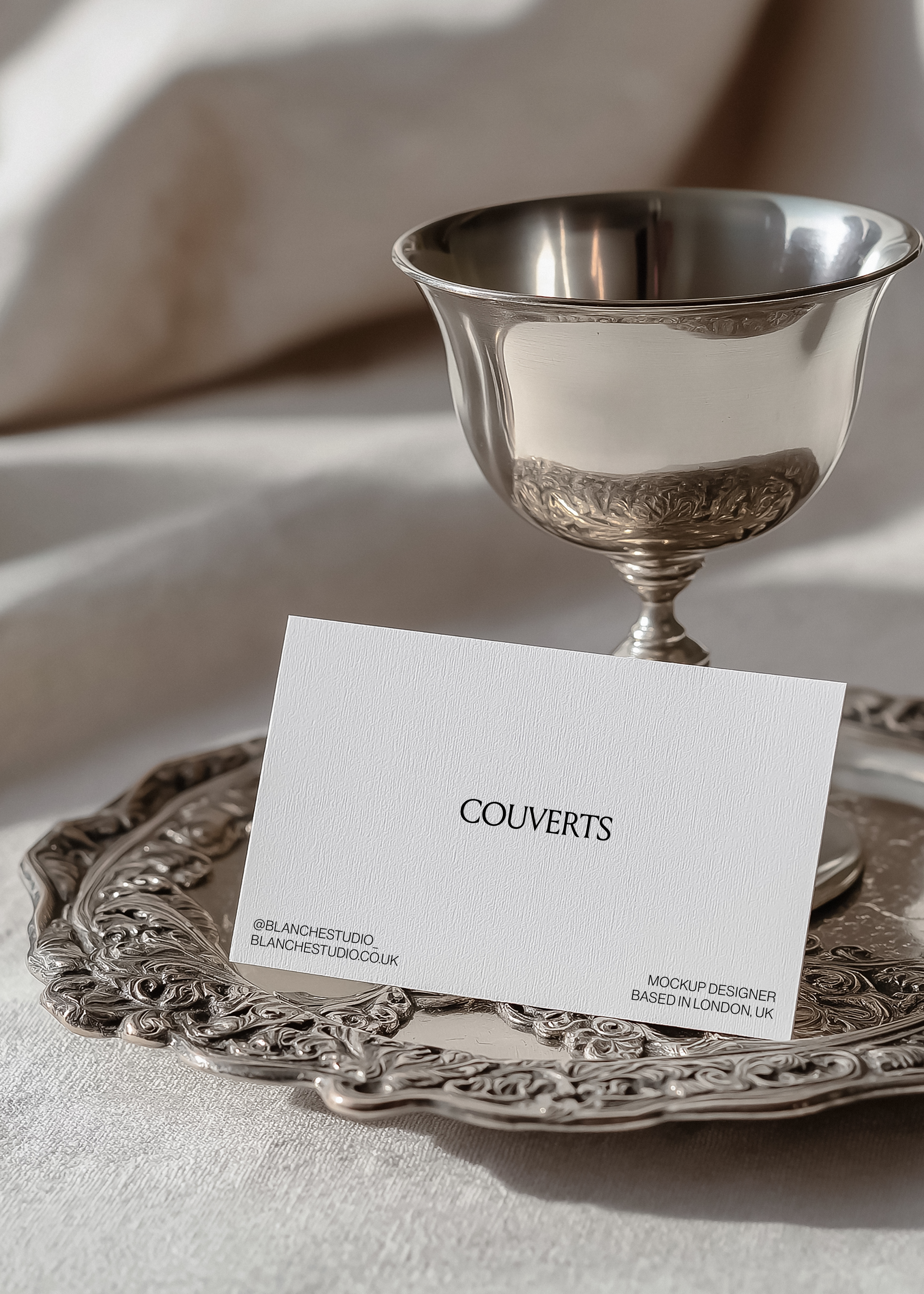 Business card mockup propped against ornate silver goblet on engraved silver tray, ivory linen backdrop, vintage editorial styling