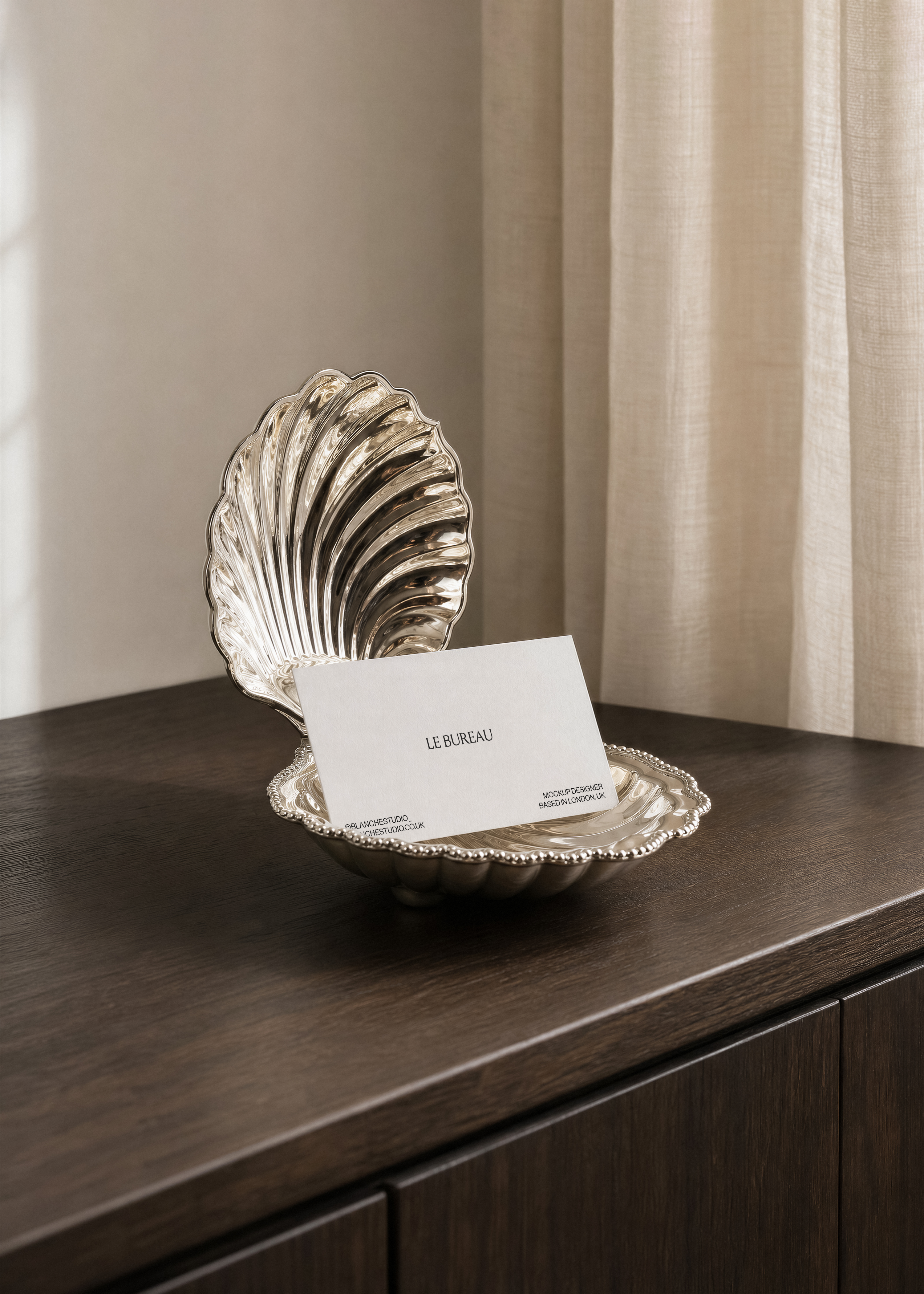 Business card mockup standing upright in silver scallop shell dish on dark wood cabinet, sheer cream curtain behind