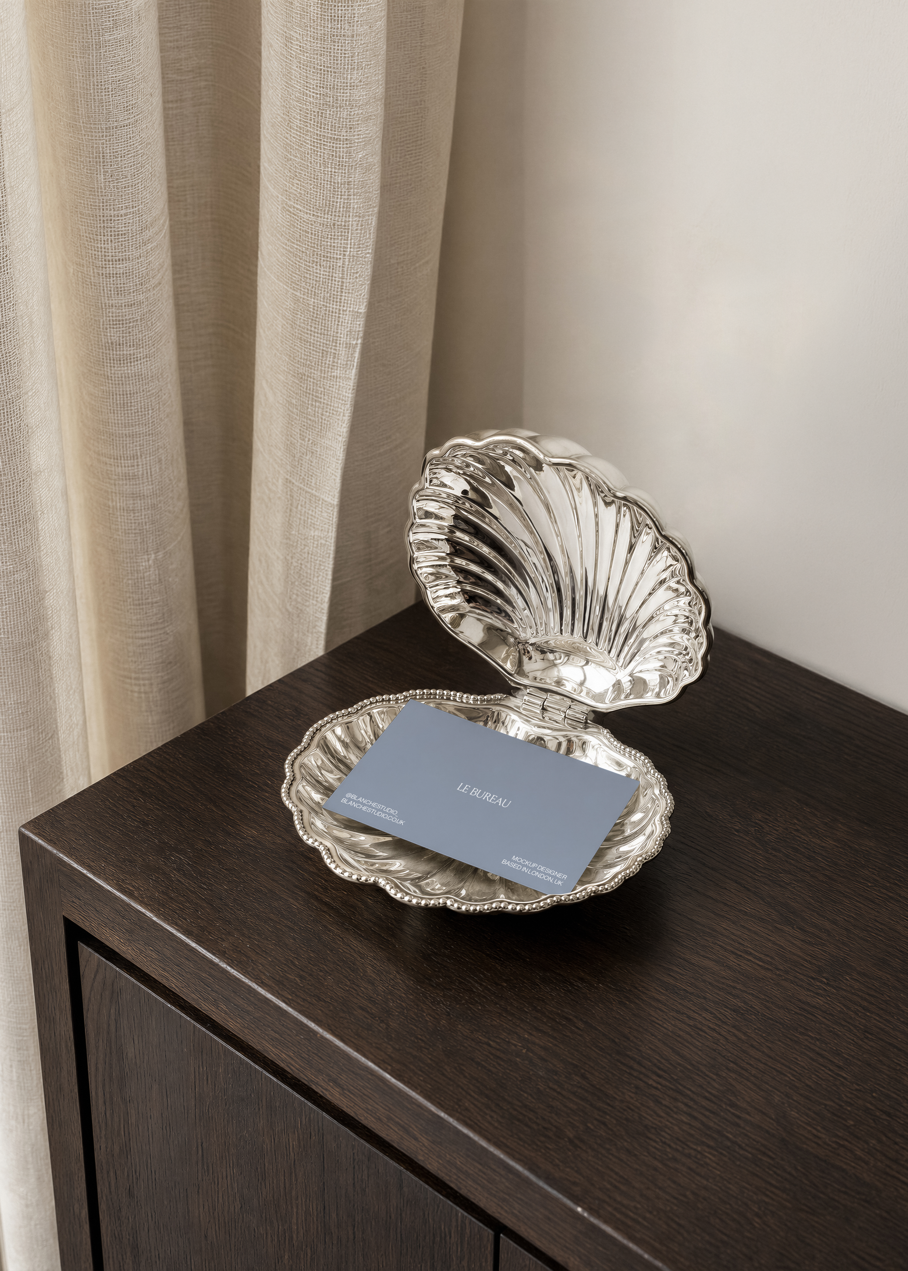 Dusty blue business card mockup with white logo inside beaded silver shell dish on dark wood, cream wall behind