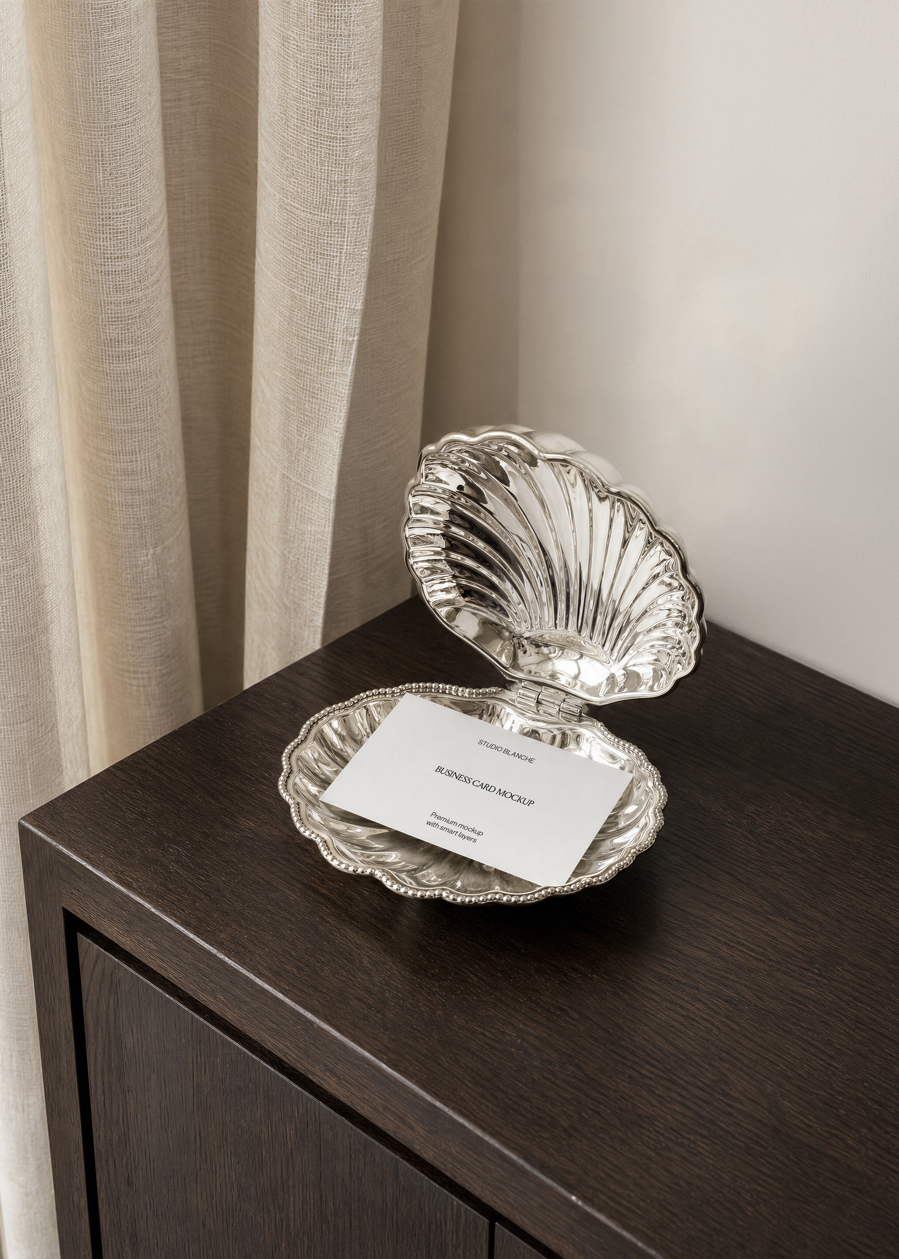 Business card mockup lying in open silver scallop shell on corner of dark oak cabinet, linen curtain, overhead view