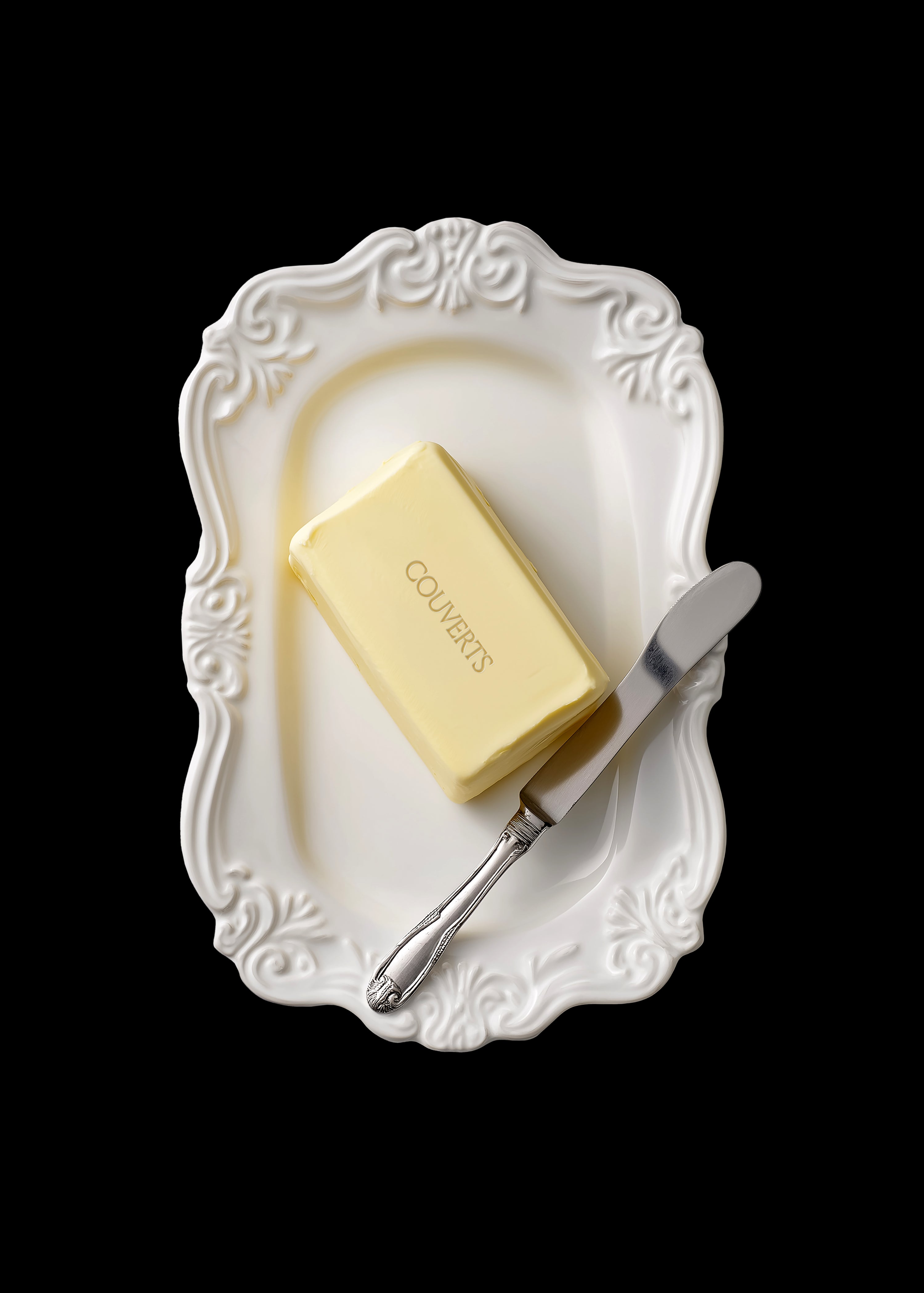 Butter block mockup on ornate white embossed ceramic dish with silver butter knife, overhead view, black background, editorial food styling