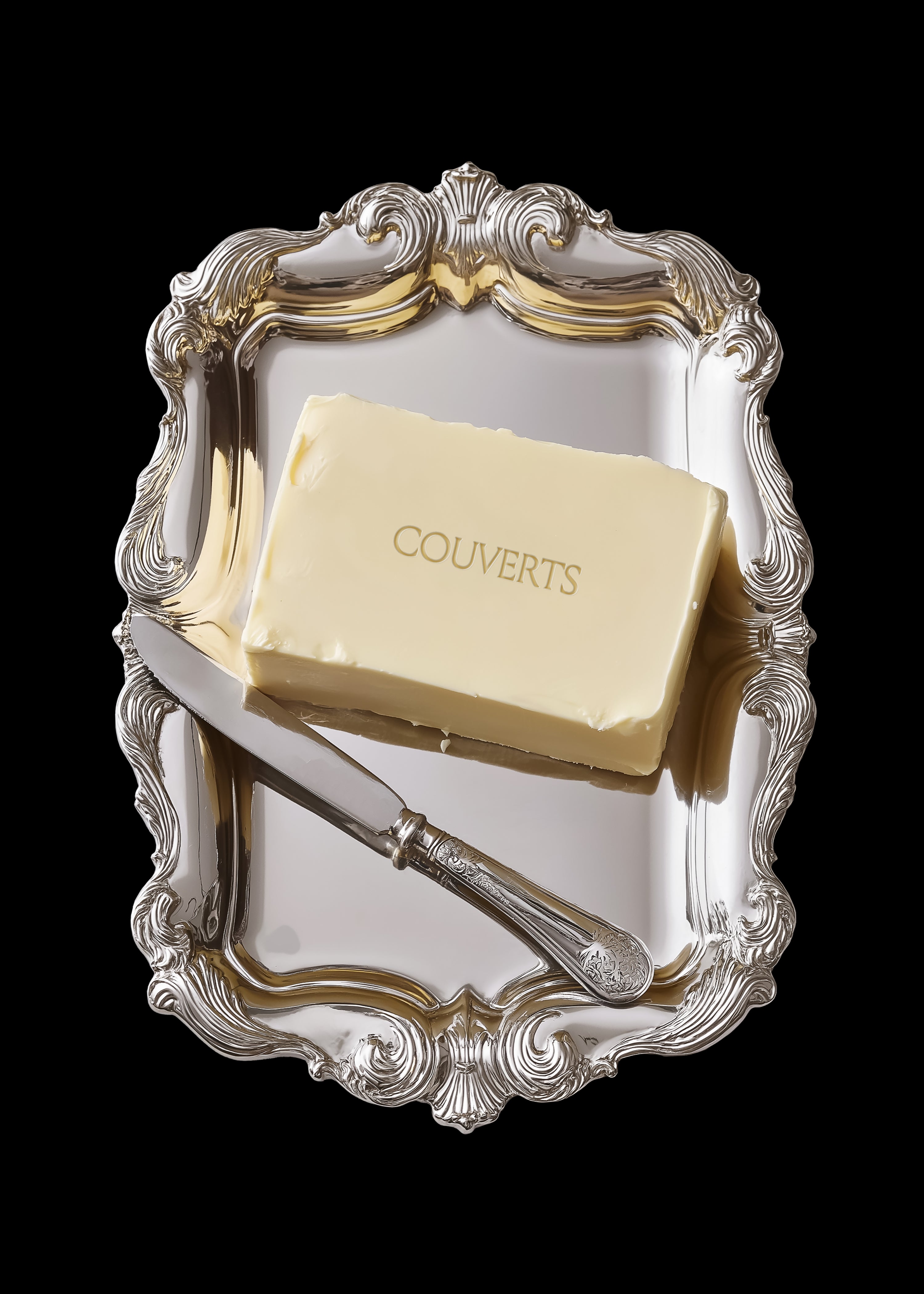 Butter block packaging mockup on ornate polished silver baroque tray with decorative butter knife, angled overhead view, black background
