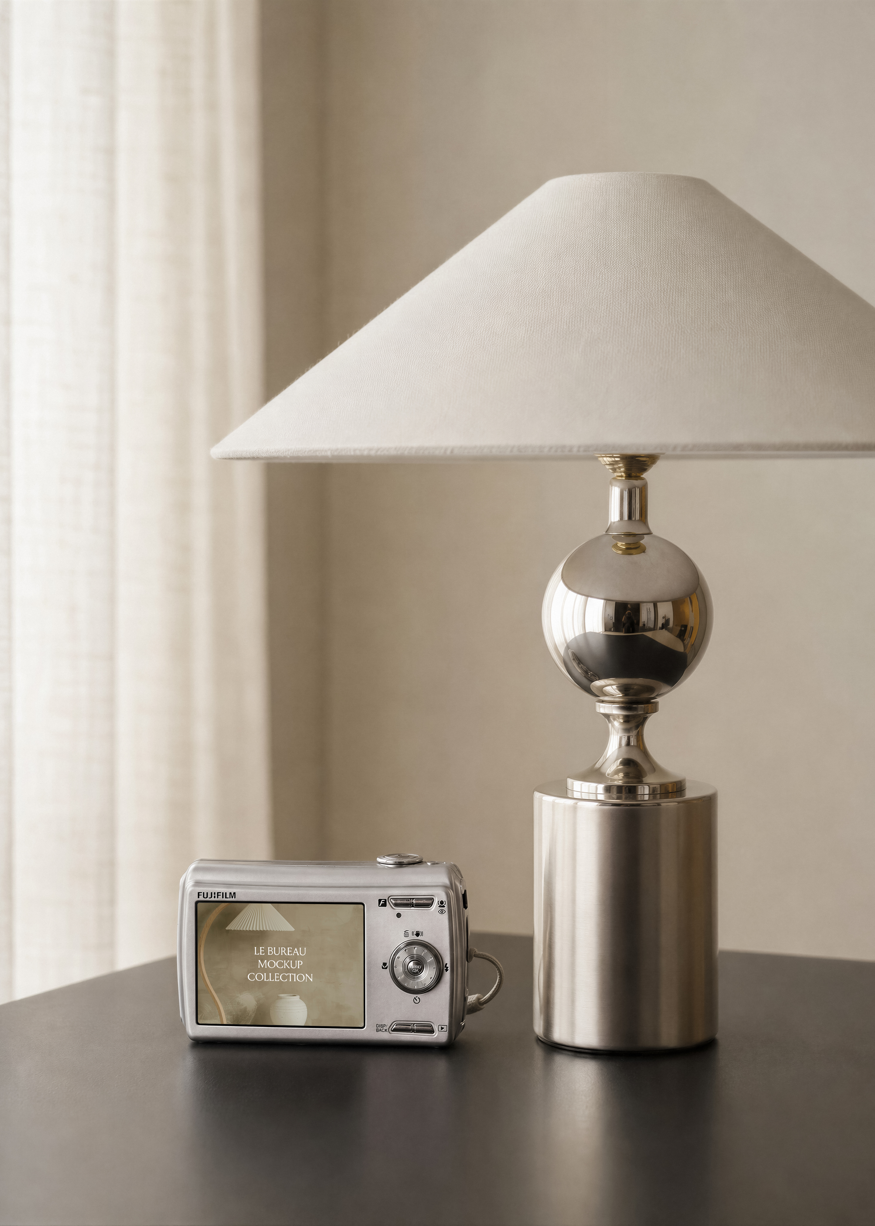 Silver digital camera mockup showing photo on screen, on dark table beside chrome ball lamp and linen curtain