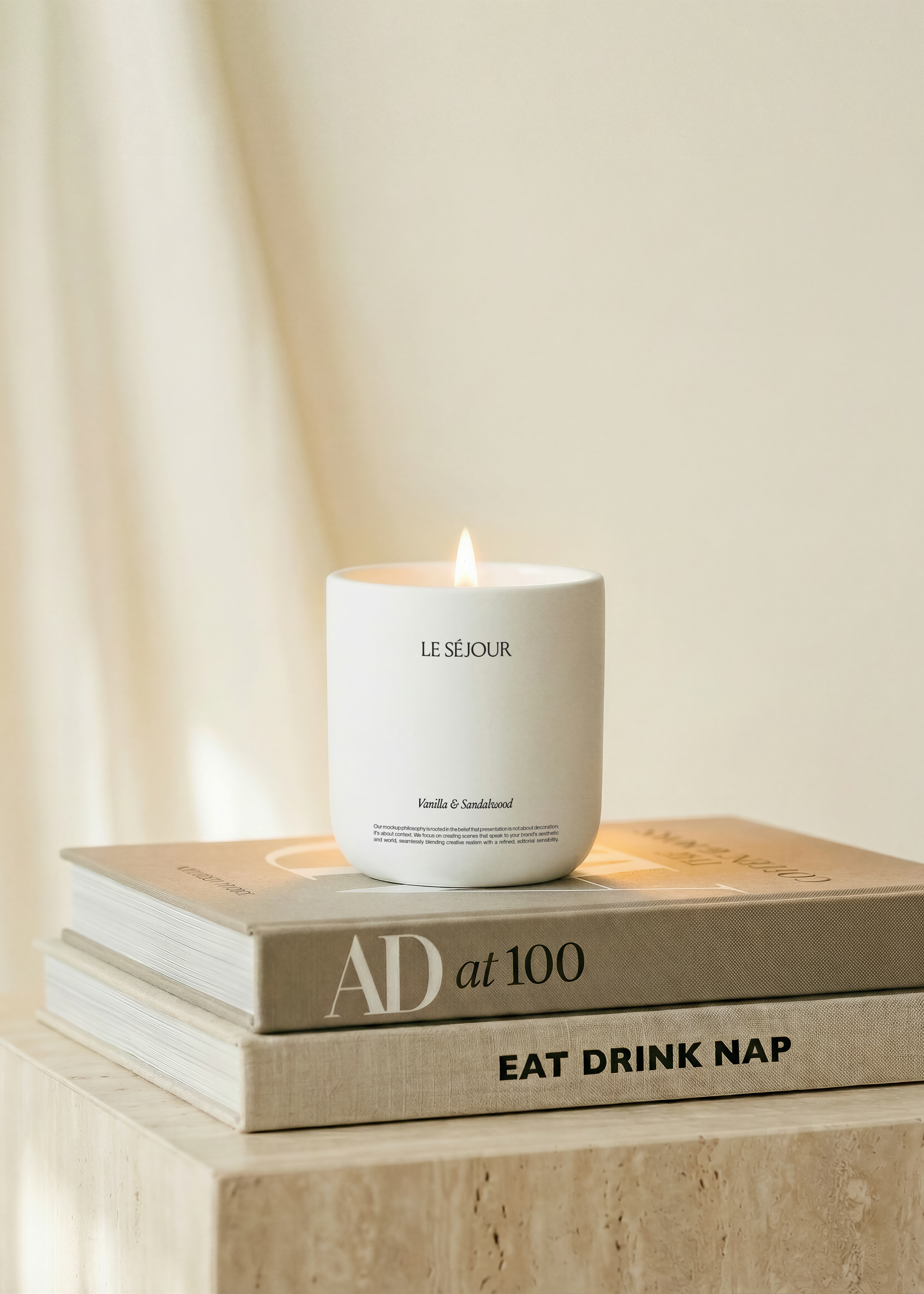Lit white ceramic candle mockup on two stacked coffee table books on a travertine plinth, cream draped curtain behind