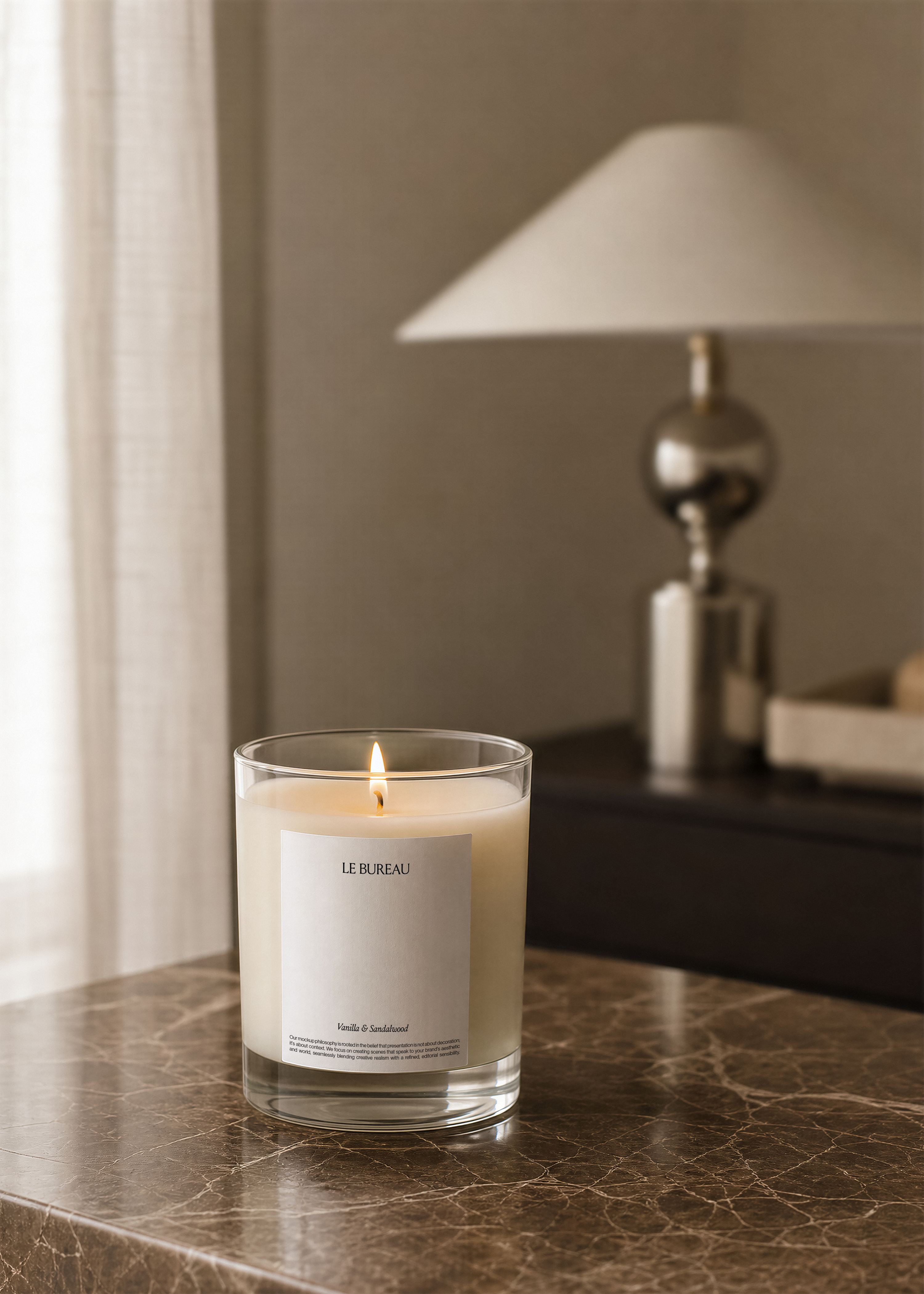 Lit glass candle jar mockup with paper label on dark marble surface, brass table lamp and curtain visible in warm blurred background