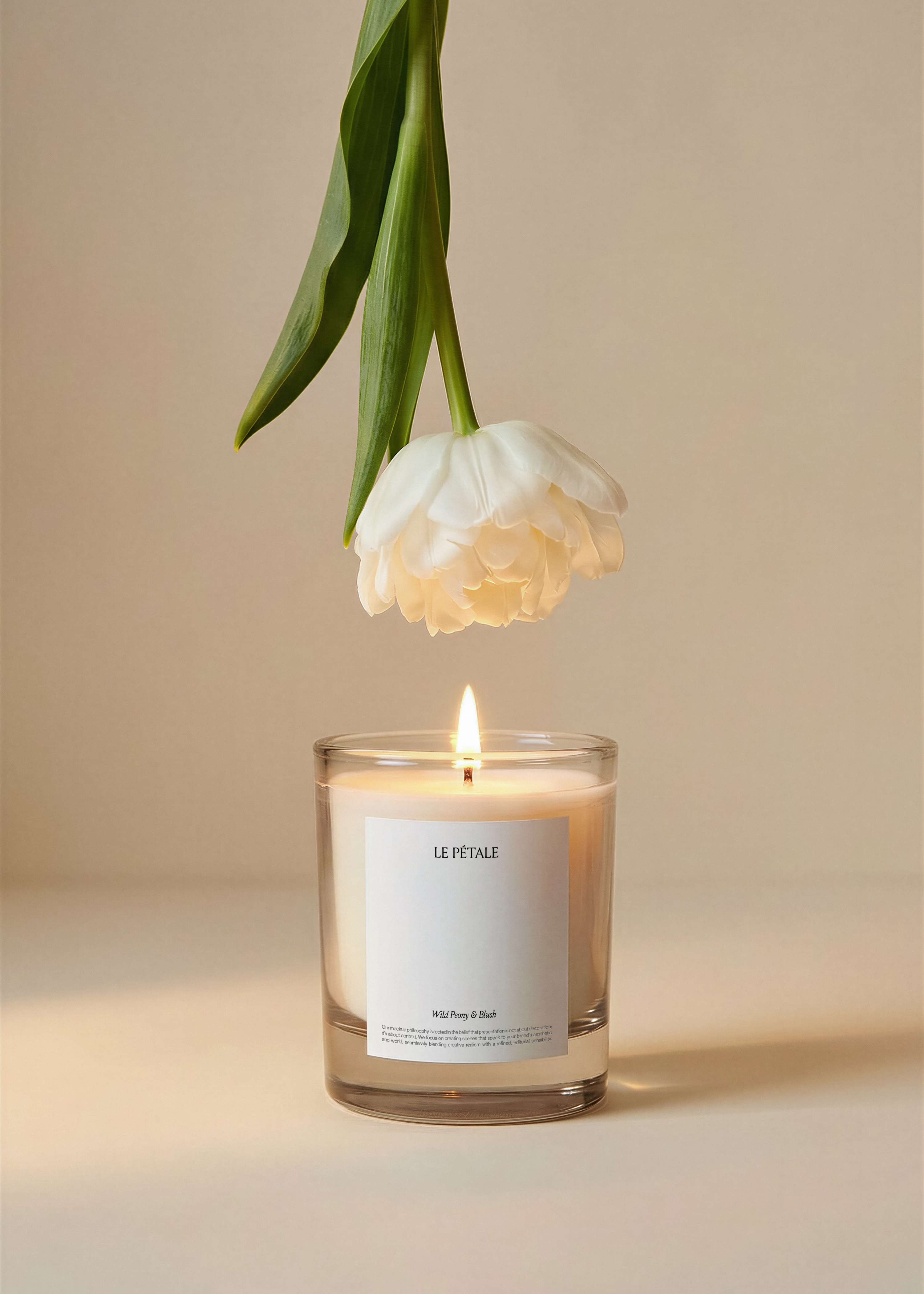 Glass candle jar mockup with white label, lit wick, white tulip suspended overhead, warm beige backdrop, botanical editorial styling