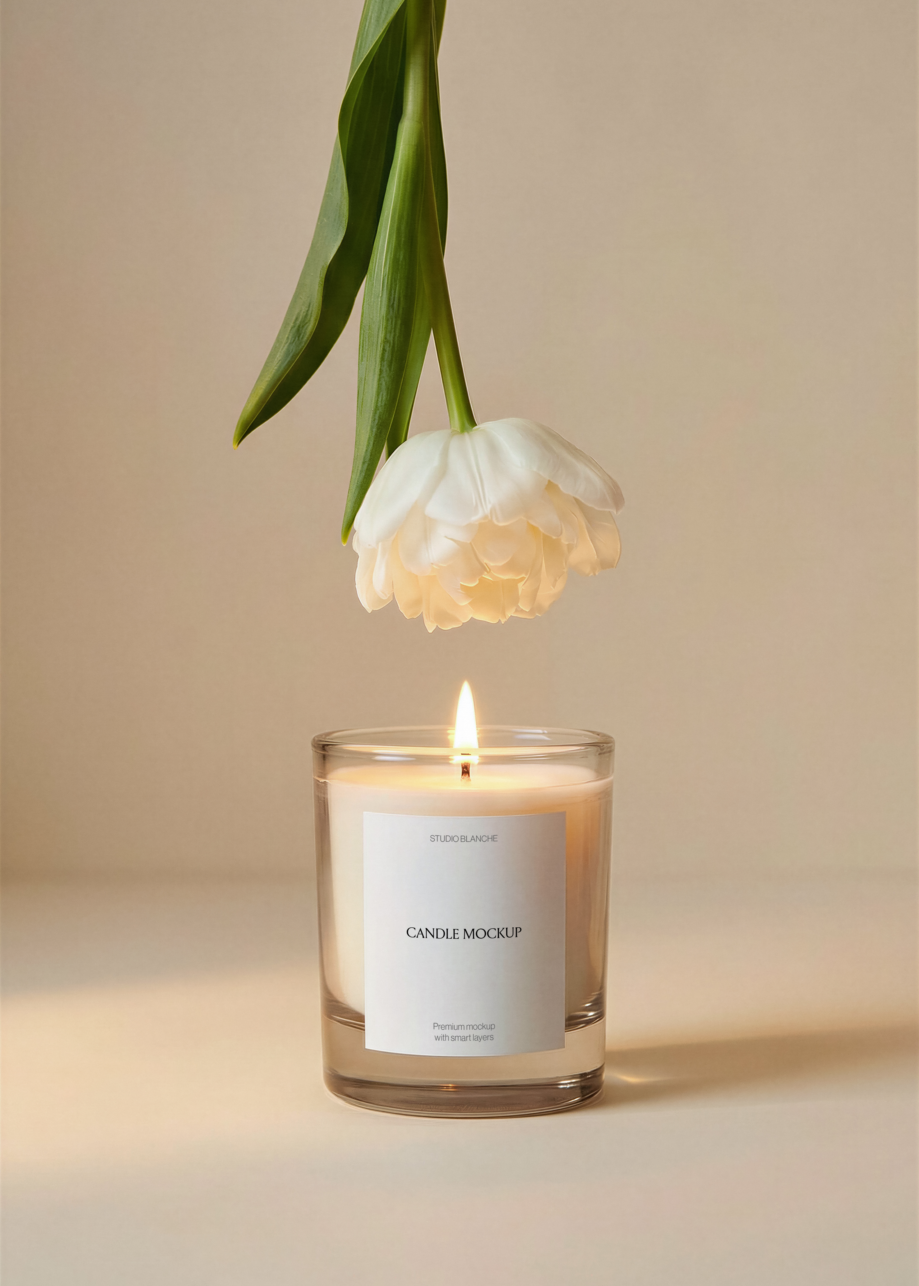 Glass candle jar mockup showing label placement area, white tulip prop, soft candlelight, neutral sand background, studio product scene