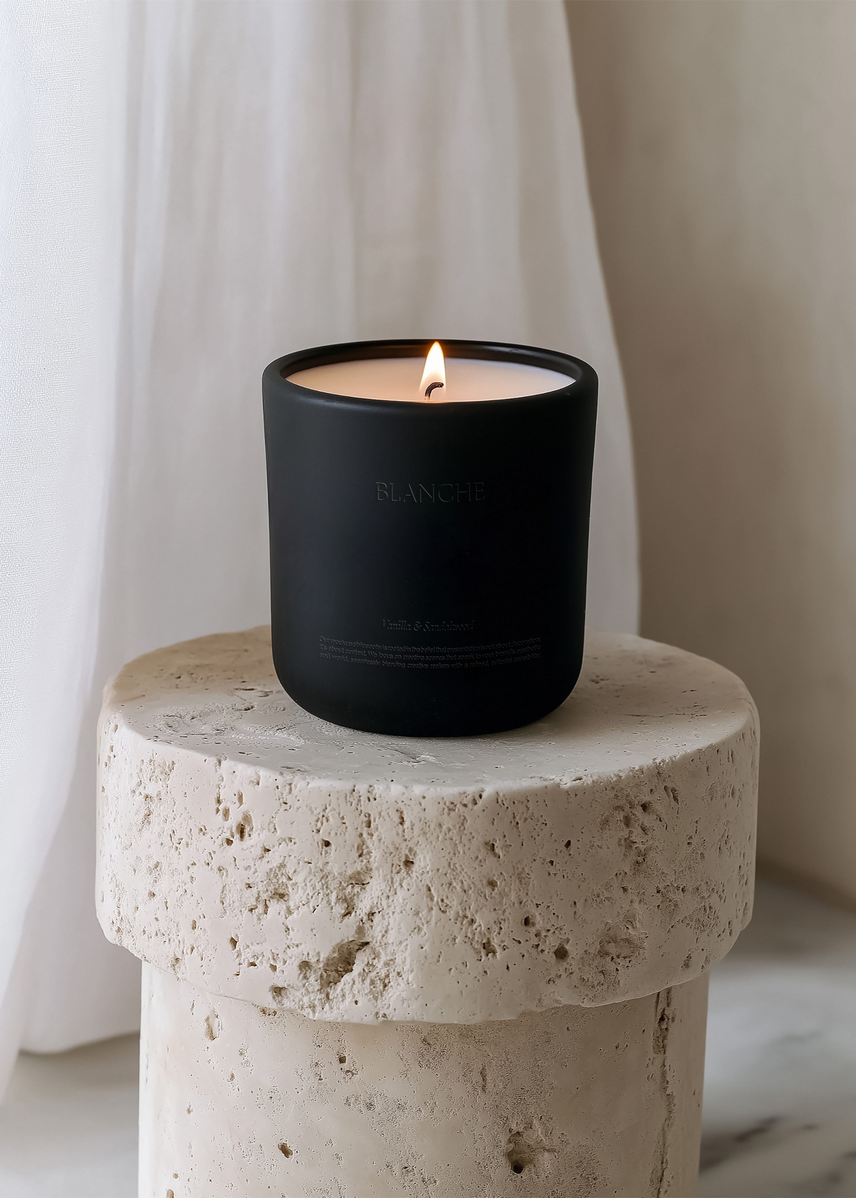 Matte black candle jar with lit flame on rough travertine cylindrical plinth, sheer white curtain backdrop