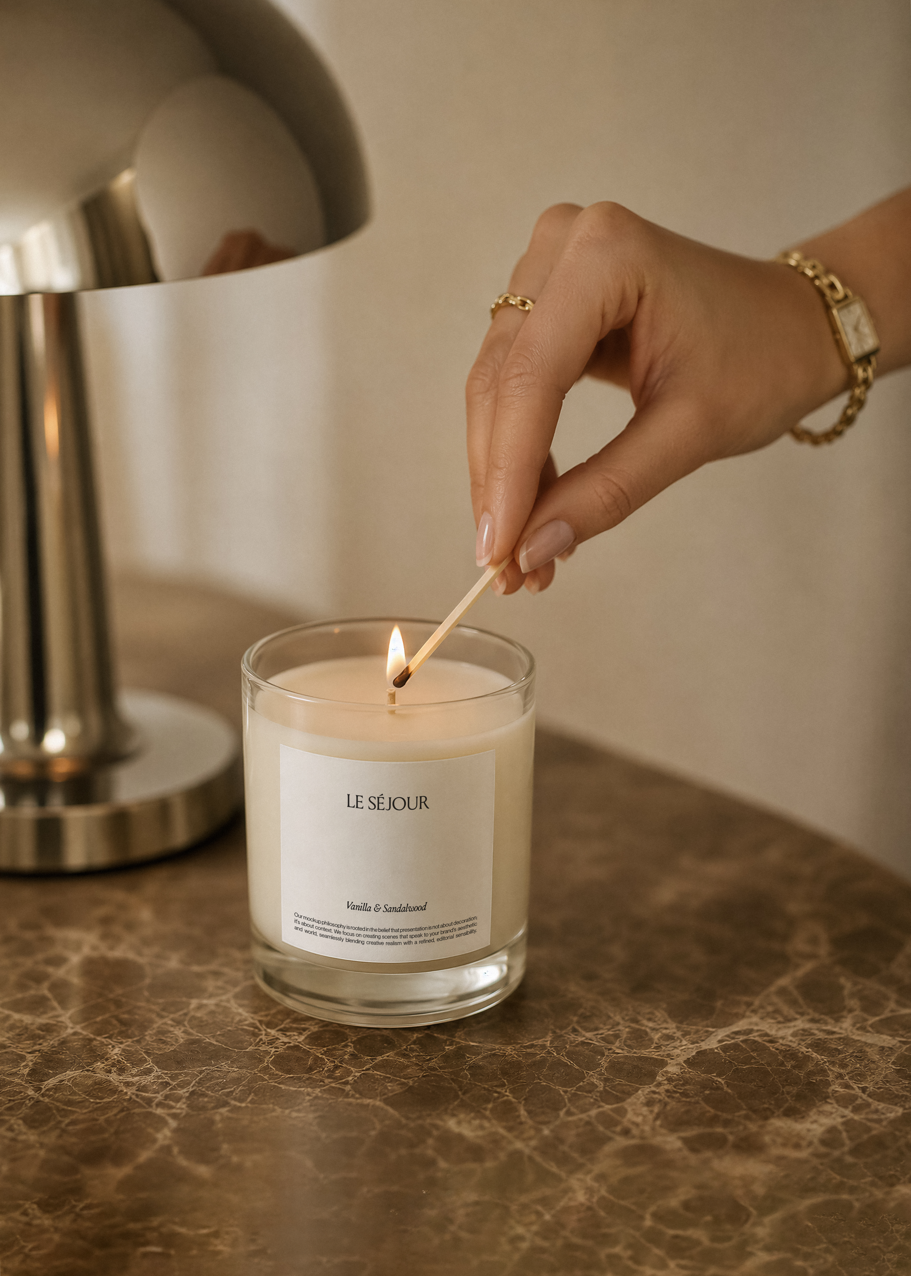 Glass candle jar mockup with white label on brown marble, a hand with gold watch lighting the wick, chrome lamp beside