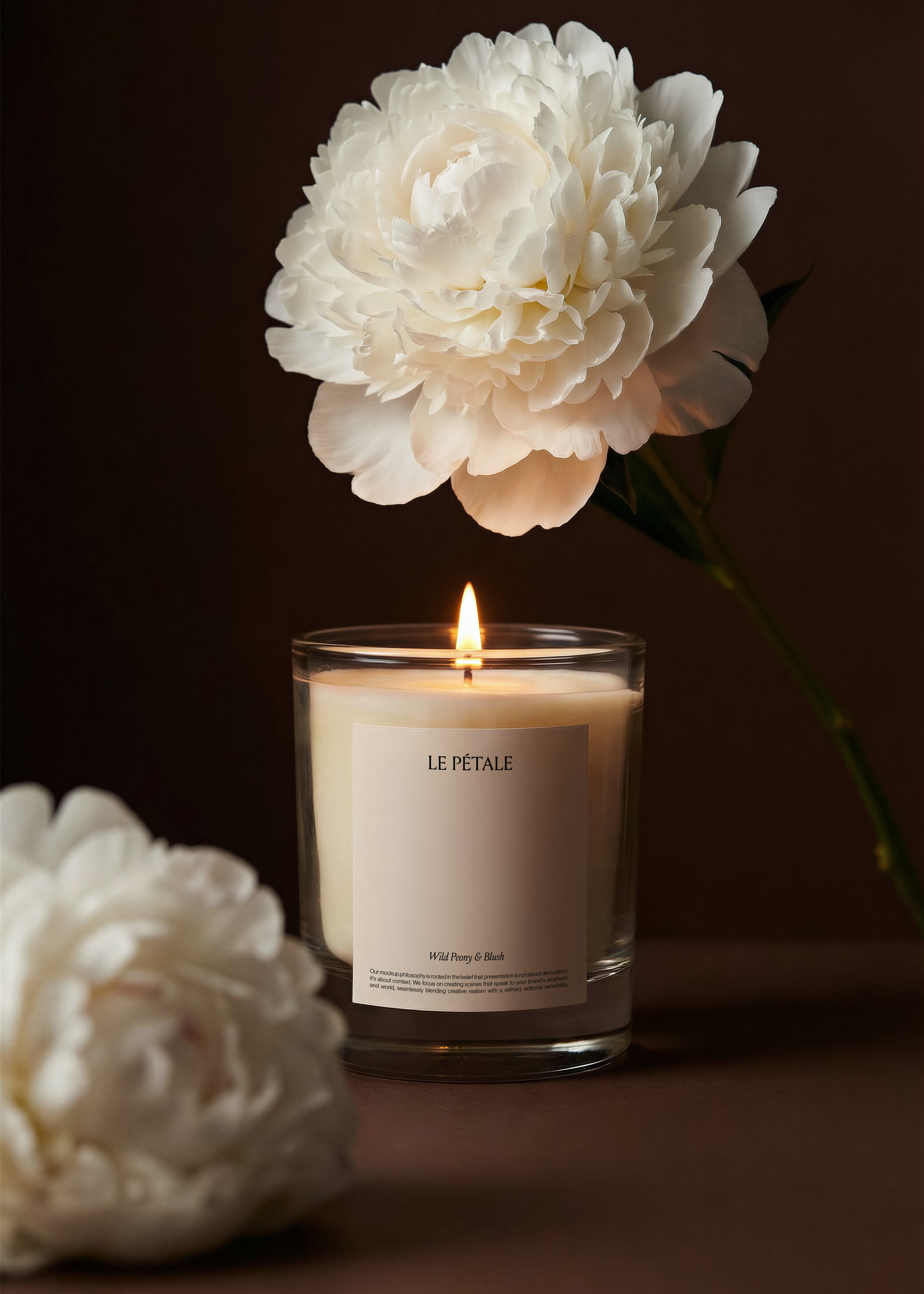 Glass candle jar mockup with white label, lit wick, large white peony overhead, fallen peony foreground, deep brown backdrop